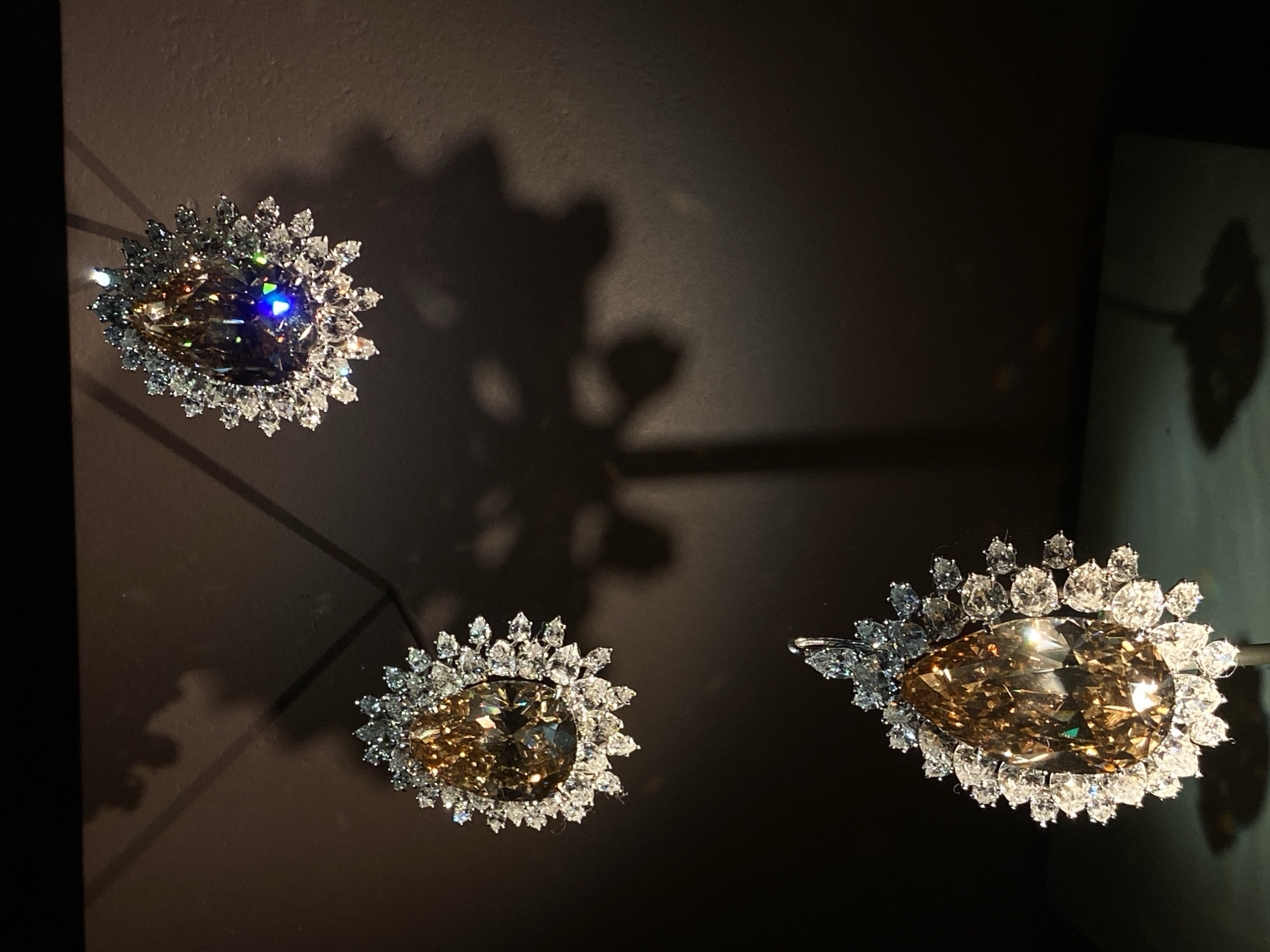 Three large, pear-shaped diamonds, each surrounded by smaller white diamonds, are displayed against a dark, textured background. The diamonds appear to be a rich amber or yellow hue, with one of them reflecting a bright blue light. Shadows of the diamonds are cast on the wall behind them, creating an artistic effect. The bottom diamond is the most prominent and detailed, showing a delicate setting. The overall impression is one of opulent display, likely in a museum or high-end jewelry exhibition. This scene could be located in Washington, D.C., potentially at the Smithsonian National Museum of Natural History, which houses a significant collection of precious gems, including large diamonds.