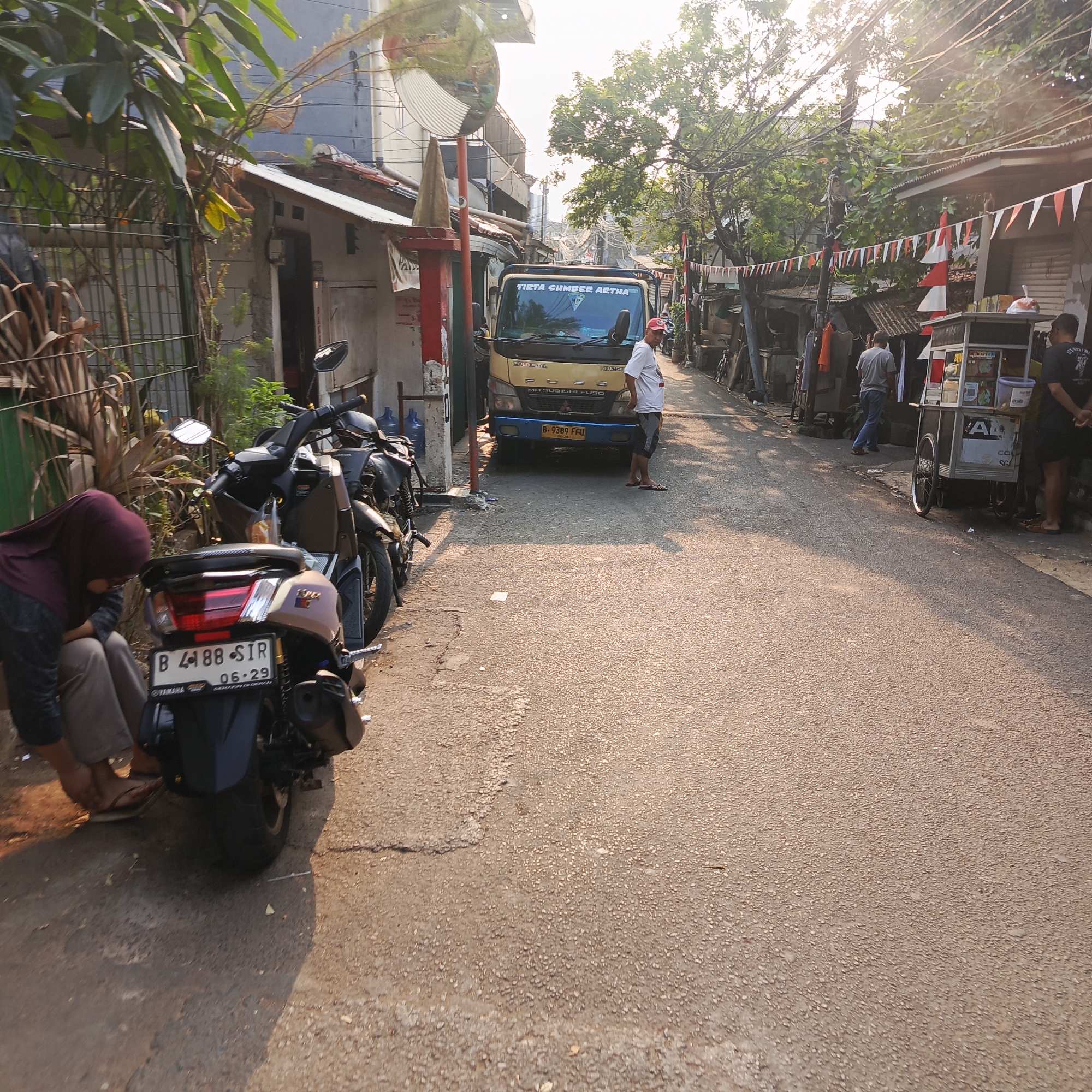 This image depicts a narrow street in South Jakarta, Indonesia, likely an alleyway or residential road given the close proximity of buildings and overhead wires. The scene is bathed in late afternoon sunlight, casting long shadows.
In the foreground, a woman wearing a maroon hijab and grey pants is squatting next to a light brown scooter, appearing to adjust her footwear. The scooter has a license plate reading "B 4188 SIR" and the date "06-29". Adjacent to her are two other motorcycles parked on the side of the road.
Further down the street, a yellow Mitsubishi Fuso truck is parked, with its headlights on and a sign on the windshield that reads "TETA SUMBER ASTHA". A man in a white t-shirt and shorts stands next to the truck, facing away from the camera.
To the right of the truck, a food cart with a blue and white exterior is set up. Two men are visible near the cart; one is wearing a black t-shirt and shorts, facing away from the camera, while another person in a grey shirt and blue jeans is walking away from the cart further down the street.
The buildings lining the street appear to be modest, some with corrugated metal roofs. Red and white pennant flags are strung across the street, suggesting a local celebration or national holiday. Trees with dense foliage are visible, and a convex mirror is mounted on a pole near the truck, likely to improve visibility around the bend. The road surface is asphalt and shows some signs of wear and tear.
