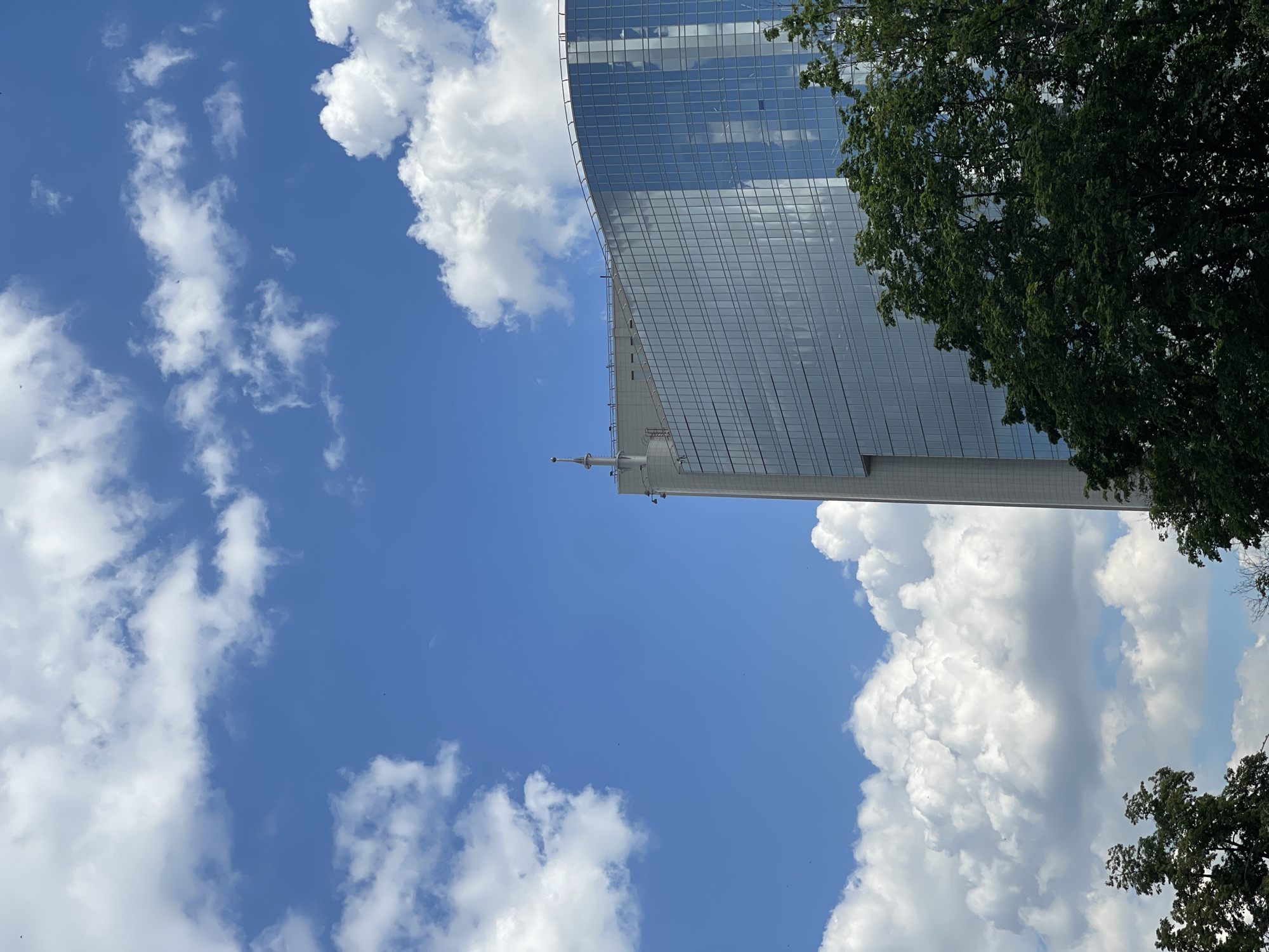 The image captures a modern glass-clad skyscraper standing tall against a bright blue sky with scattered white cumulus clouds. The building's reflective facade mirrors the sky and clouds, creating a dynamic interplay of light and shadow. Its design features a distinctive curve, giving it a sleek appearance. Towards the upper portion of the structure, a slender antenna or spire is visible, adding to its architectural detailing.
Dark green foliage from trees partially frames the right side of the image, overlapping with the building. The sky dominates the left and central parts of the frame, showcasing clear blue expanses interspersed with fluffy white clouds. Several tiny, dark specks, likely birds, are visible against the bright sky.
The scene suggests a clear, sunny day, most likely during midday or early afternoon, given the strong illumination. There is no human activity evident, and the image focuses primarily on the architectural structure and natural elements. No visible text is present. The provided context indicates the location is Moscow, Russia.