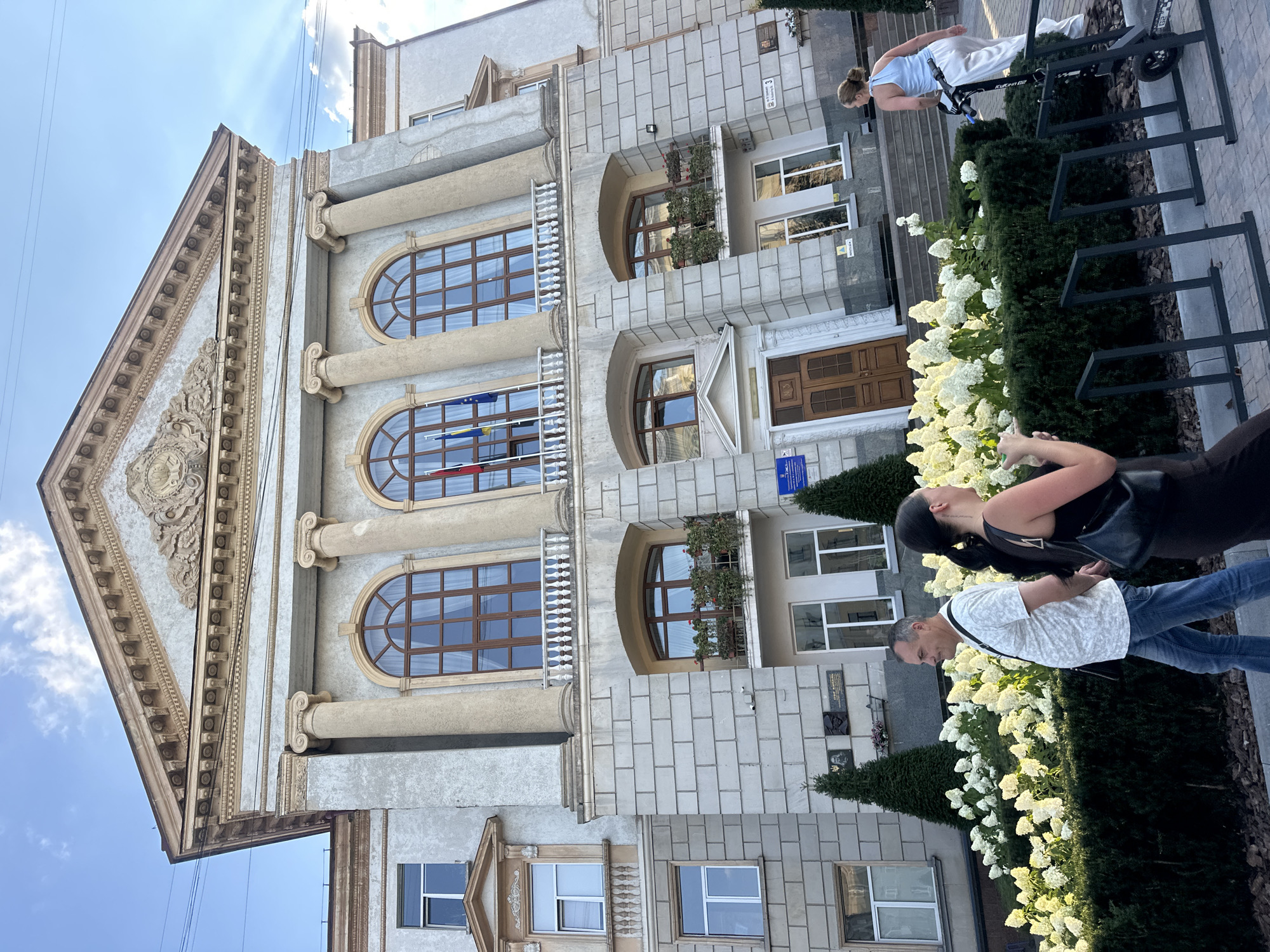 In the city of Ternopil, Ukraine, a three-story building with classical architectural elements stands under a bright, partly cloudy sky. The building features a pediment adorned with a decorative relief and large, arched windows with wooden frames and white balconies. Three prominent Corinthian columns are visible on the second story. In front of the building, a line of lush green bushes and white hydrangeas creates a vibrant border. Two individuals are walking on the sidewalk in the foreground. A man in a white t-shirt and blue jeans is walking to the left, and a woman in a dark dress is walking to the right, holding a black purse. Further to the right, a woman in a light blue top and white pants is riding a scooter. The sidewalk is lined with a bike rack, and a few trees are planted near the building. The scene captures a bright, sunny day in a public urban space.