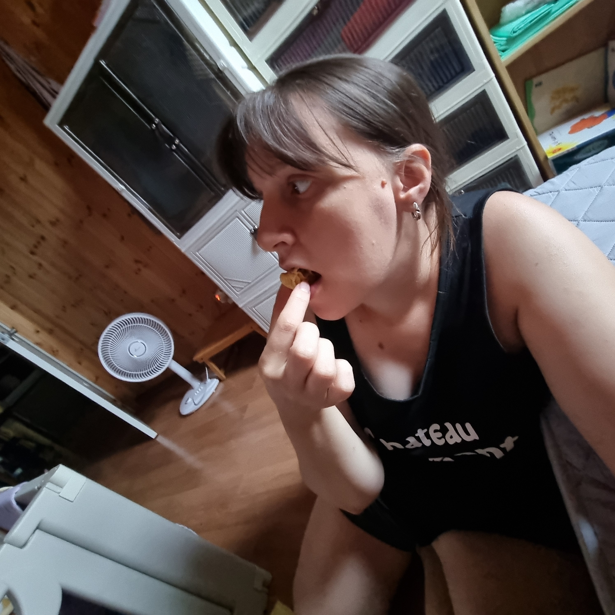 A woman is in a room, possibly a bedroom or living area, with wooden floors and built-in white cabinetry. She is wearing a black tank top with white text that reads "chateau". She is holding a small, light-colored food item to her mouth with her right index finger and thumb, appearing to be eating it. Her hair is dark brown and styled with bangs. In the background, a white oscillating fan is visible on the floor. To the left, part of a light-colored storage unit or appliance is visible. To the right, a quilted surface, possibly a bed or couch, is present. A shelf with books and green plastic items is also visible in the background. The overall lighting suggests an indoor setting. No information about the location in Dunnae-myeon, South Korea is discernible from the image.
