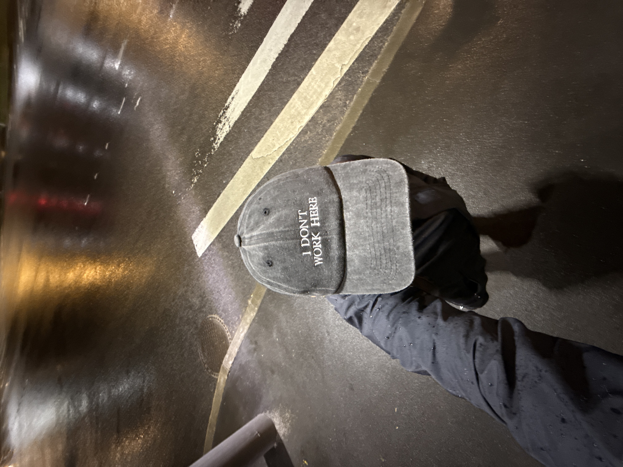 A grey baseball cap with the phrase "I DON'T WORK HERE" embroidered in white lies on a wet, dark pavement with painted white stripes and street lighting reflections. A person wearing a dark, possibly rain-slicked jacket stands nearby. The location is Moscow, Russia.