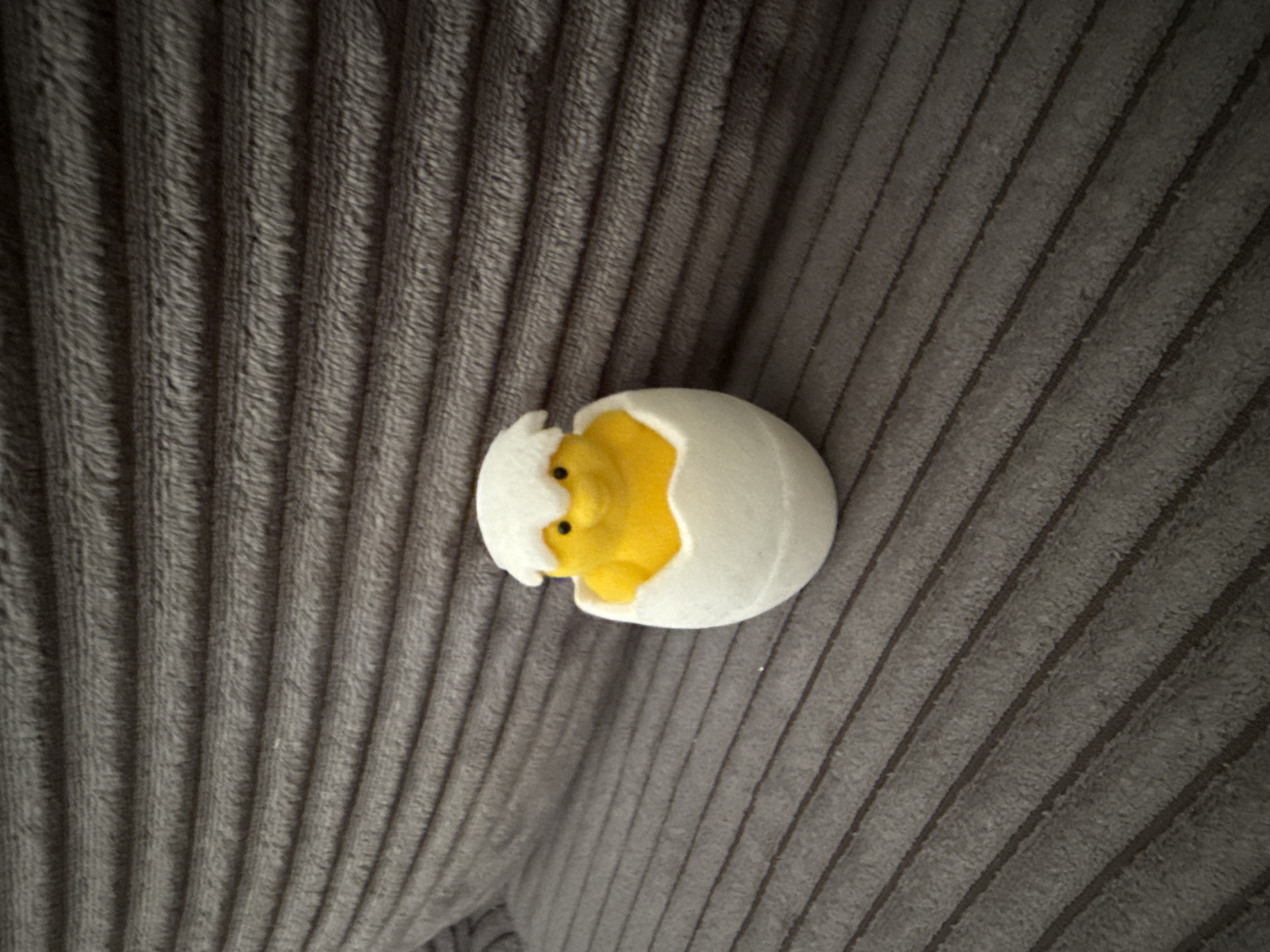 A close-up shot depicts a yellow chick toy emerging from a cracked white eggshell. The toy is situated on a gray corduroy surface. The location is not discernible from the visual information provided.