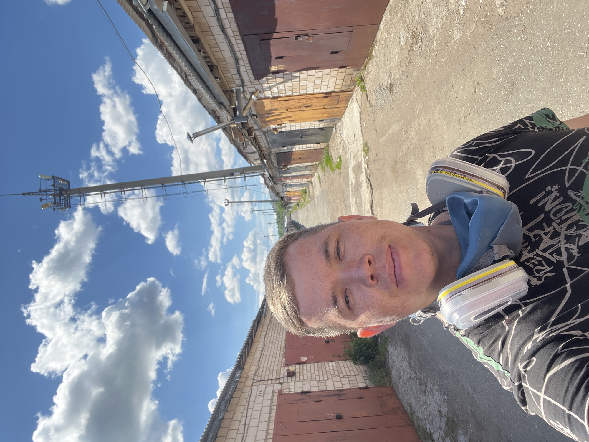 A man wearing a black t-shirt with a white geometric pattern and a respirator mask around his neck stands in front of a row of brick garages. The garages have corrugated metal doors of various colors, mostly reddish-brown. A tall communications tower is visible in the background, along with a bright blue sky with scattered white clouds. The ground is paved with asphalt or gravel. The location is identified as Veteran-2, Russia.