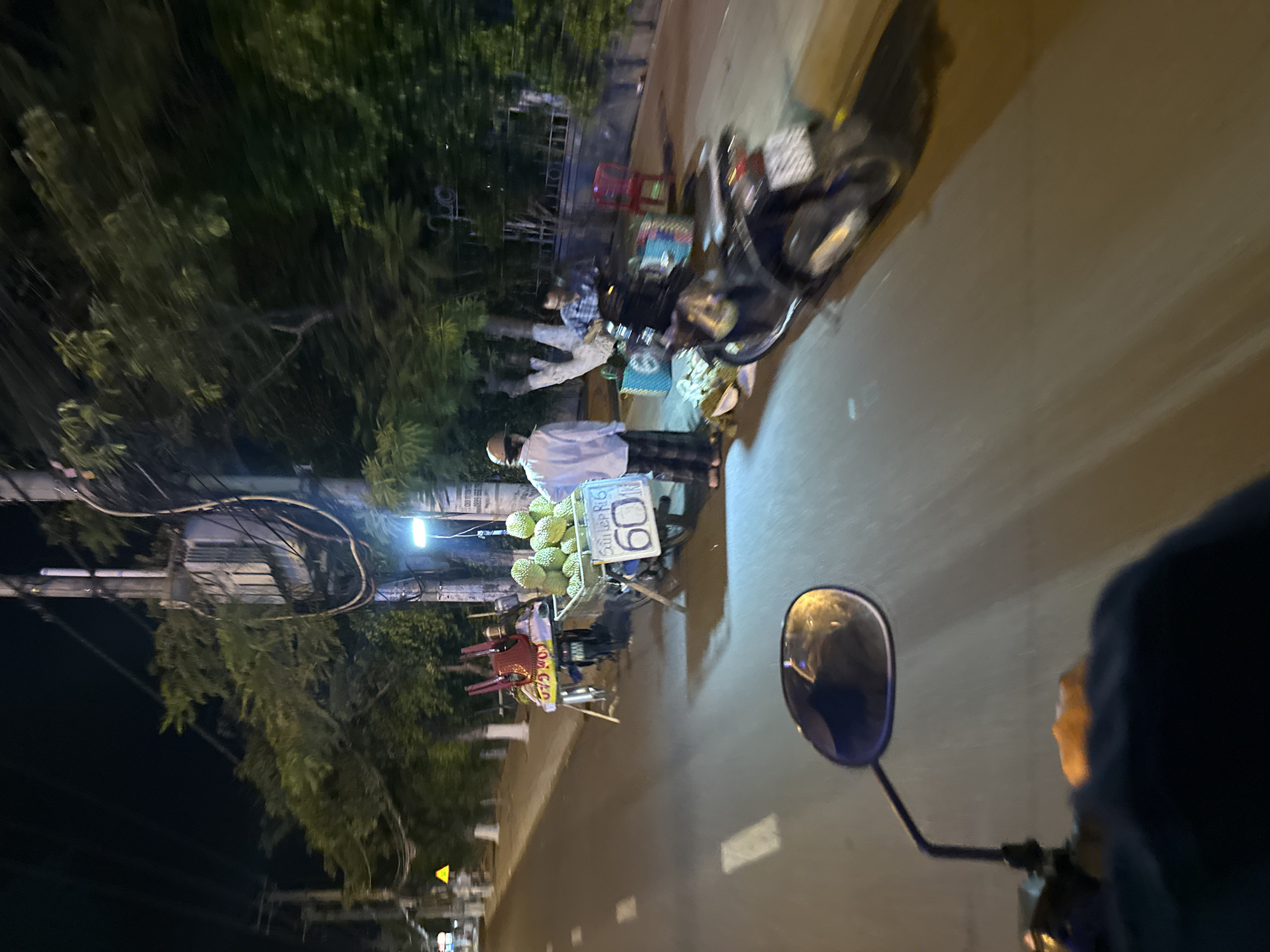 The image captures a nighttime scene in Bac Nha Trang Ward, Vietnam, from the perspective of a person riding a motorbike. A street vendor selling durian is visible on the side of the road. The vendor is wearing a white shirt and hat, with durians displayed on a cart with a sign indicating the price. Other items like plastic chairs are also visible in the vicinity of the vendor. A second person can be seen sitting near the cart as well. The background shows buildings and trees. Street lights illuminate the scene, and the motorbike's mirror is visible on the right side of the frame.