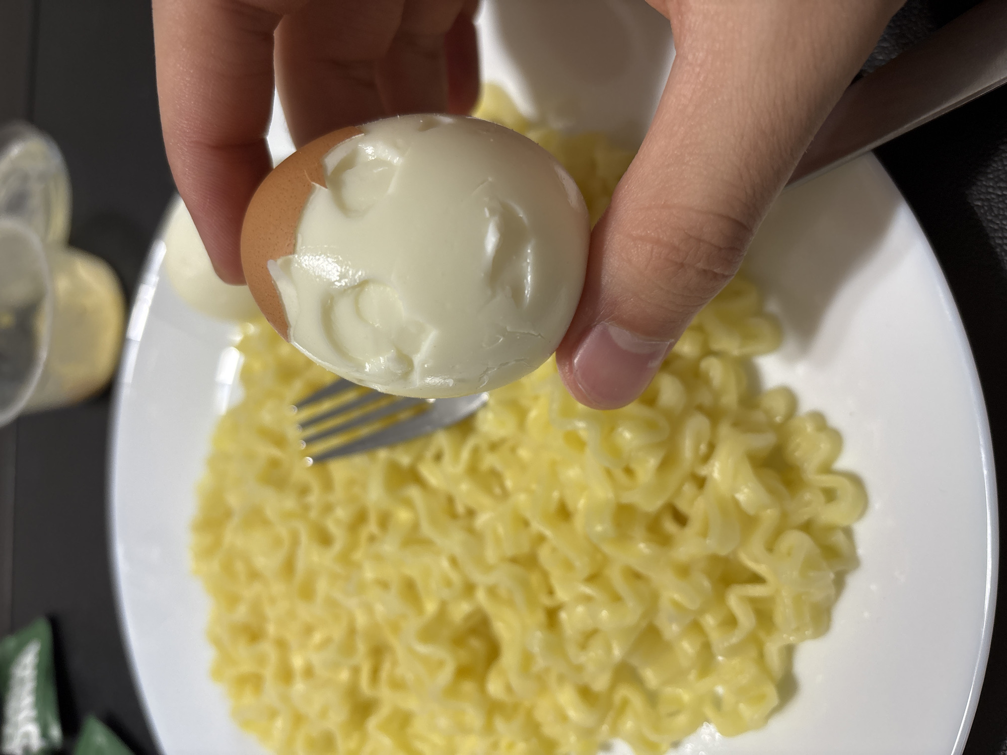 A person is holding a partially peeled hard-boiled egg over a plate of yellow pasta with a fork resting on the plate. Another hard-boiled egg is visible on the plate. The setting appears to be indoors. The city of Franklin Township, United States of America is not explicitly discernible in the image.