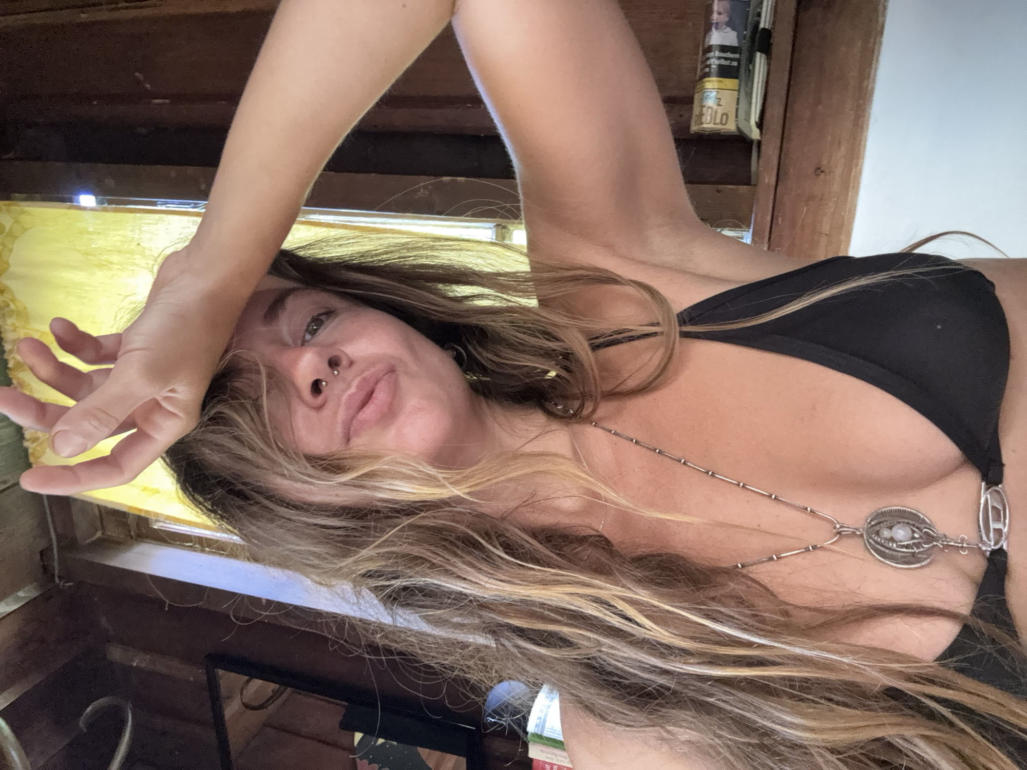 A young woman with long, blonde-streaked brown hair is taking a selfie in a dimly lit room. Her hair is spread across her face and shoulders, obscuring parts of her features. She is wearing a black bikini top with a silver necklace that has an eye-shaped pendant. Her right arm is raised, with her hand resting on her forehead, as if she is shielding her eyes from the sun. Her left hand is extended upwards, with fingers splayed.
The background suggests an indoor setting, possibly a rustic cabin or an old building, with wooden beams and shelves visible. There's a yellow-tinted window or light source to her left, casting a warm glow. On a shelf behind her, a product with text is visible, including the word "EDLO" and a partial image of a person. The overall mood is relaxed and intimate, with the woman appearing to be enjoying a moment of repose. The time of day and weather are not discernible from the image.