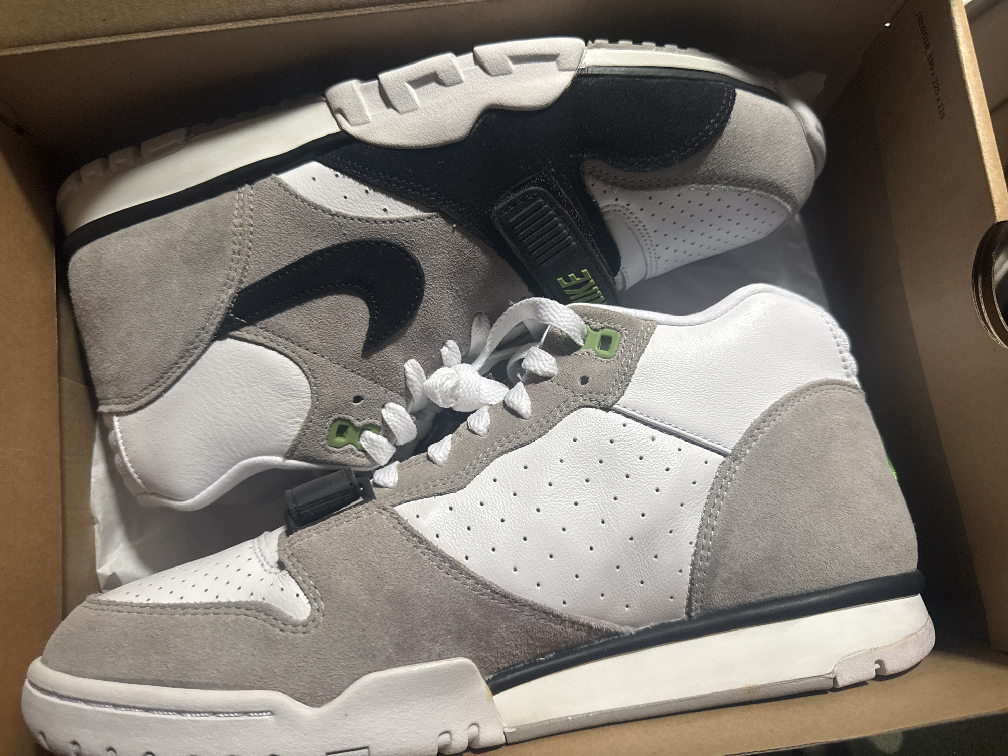 The image shows a pair of Nike Air Trainer 1 shoes inside a cardboard box. The shoes are mostly white and gray with black accents. The white parts appear to be perforated leather, while the gray areas seem to be suede. One shoe is slightly on top of the other. The background consists of the interior of the box and some crumpled white paper. There is no indication of Barcelona, Spain, or any other location, people, or actions within the image.