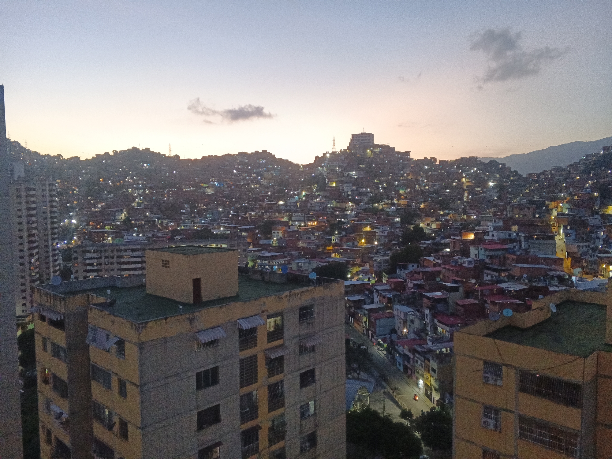 This is an aerial photograph of a densely populated hillside neighborhood in Caracas, Venezuela, at dusk. The foreground features two apartment buildings, one tall and modern on the left, and a lower, older building with a distinct yellow facade and a small, block-like structure on its roof on the right. The buildings are characterized by multiple windows, some with awnings, and visible air conditioning units.
Beyond the foreground buildings, the landscape transitions into a sprawling favela that covers the hillsides. Thousands of brightly lit homes, with red and multicolored roofs, are clustered together, creating a vibrant tapestry of lights against the darkening sky. The uneven terrain of the hills is accentuated by the density of the settlement.
In the background, a larger, more structured building with a prominent antenna stands atop one of the hills. The distant mountains of Caracas are faintly visible against the twilight sky, which exhibits soft hues of purple, pink, and orange. The overall impression is one of a vast, active urban area existing in a challenging, hilly topography. No people are clearly discernible in the image.