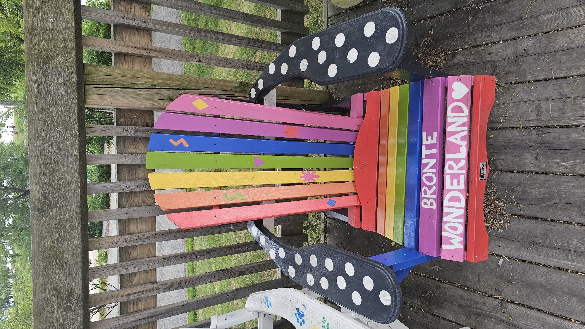 This media file features a vibrantly painted Adirondack chair with rainbow-colored slats. The chair has black armrests decorated with white polka dots. The slats of the chair are painted in the colors of the rainbow, from red to violet. On the violet and pink slats, the words "BRONTE WONDERLAND" are written vertically in white lettering. A small black and red sticker with the word "BUM" is visible on the right side of the chair. The chair is positioned on a wooden deck, with a weathered wooden fence and railing behind it. Green grass and trees are visible in the background, suggesting an outdoor setting. The lighting and shadows indicate it is daytime, likely a bright day with no obvious signs of inclement weather. There are no people clearly visible in the frame, but the setting suggests a park or garden area.