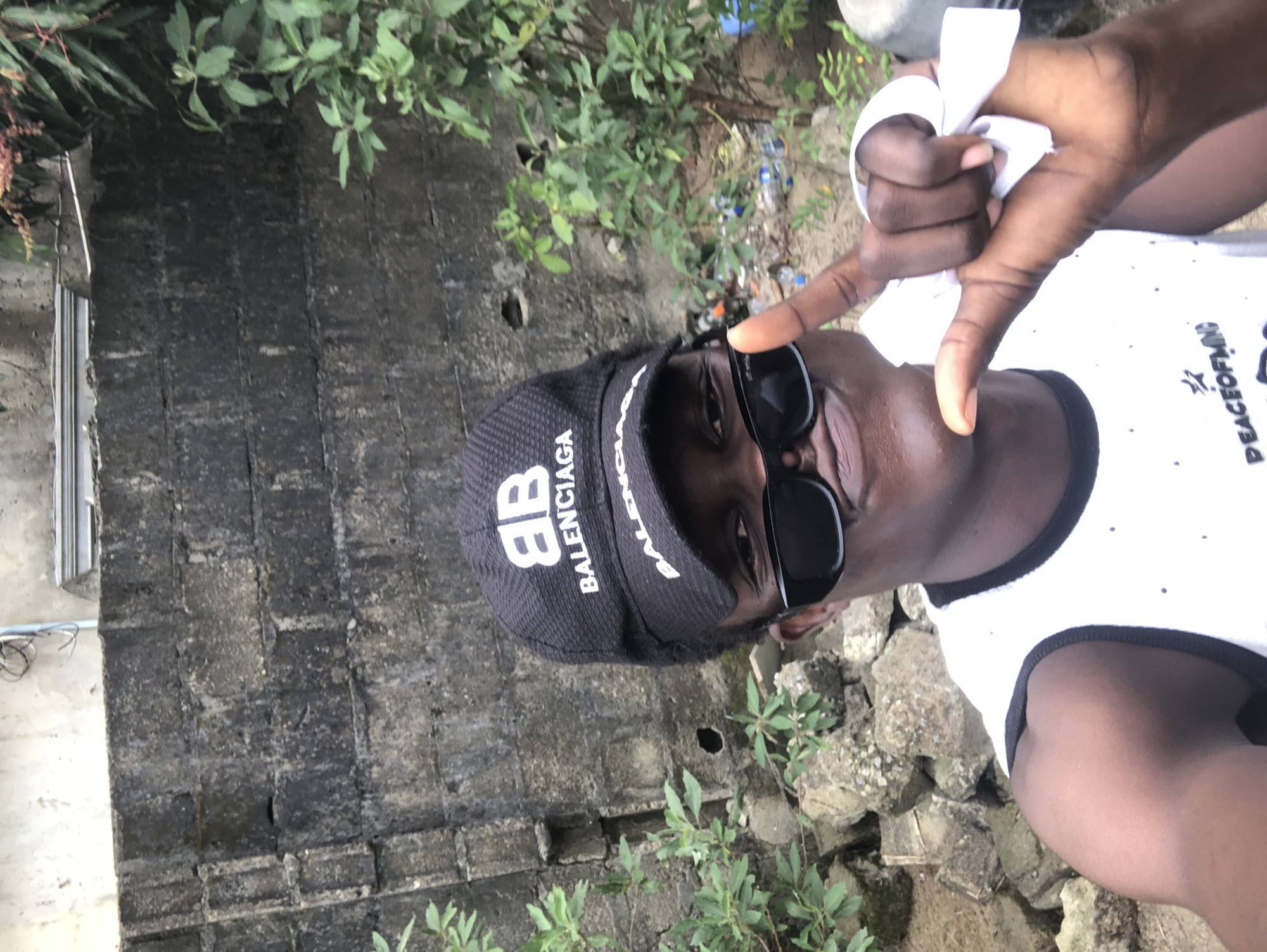 A young Black male is taking a selfie outdoors, with his face positioned in the lower right quadrant of the image. He is wearing a black baseball cap with the "BALENCIAGA" logo, black sunglasses, and a white tank top with a black trim. He is raising his right hand, showing a peace sign gesture and holding a white piece of paper. Behind him is a weathered, rough-textured concrete wall, with some greenery and scattered debris on the ground. The scene suggests an informal outdoor setting, potentially in a less maintained area of Ado, Nigeria, with the background elements indicating a possibly urban or semi-urban environment.