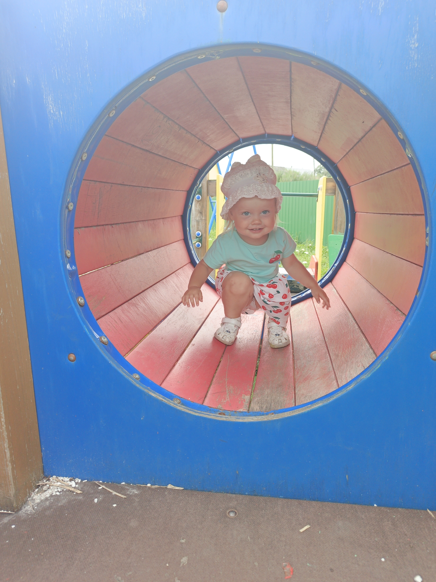 A young child wearing a pink bonnet, a light blue t-shirt with a cherry design, and white shorts with a cherry pattern is crouching inside a blue and red tunnel on a playground. The child is smiling and looking towards the camera. The tunnel is constructed of wooden planks inside and painted blue on the exterior. In the background, a green fence and some vegetation are visible. The ground in front of the tunnel is a brown, textured surface. This playground is located in the city of Medvedyovka, Russia.