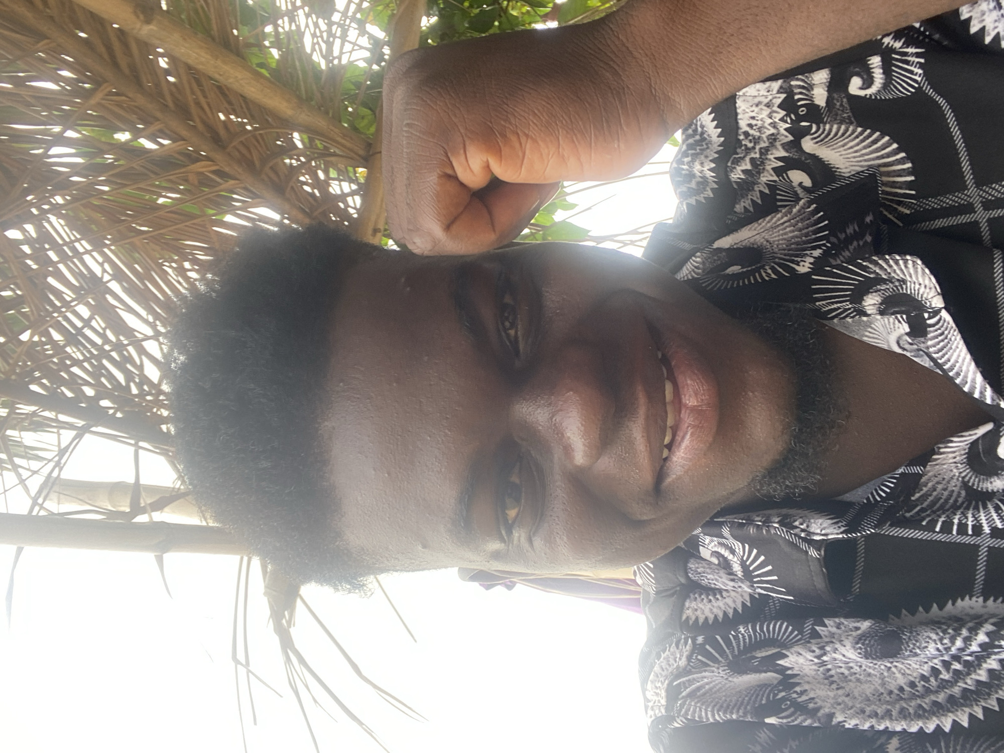 A close-up, low-angle shot depicts a dark-skinned man smiling slightly, with a fist hovering above his forehead. The background includes palm fronds and foliage, suggesting an outdoor setting. The man is wearing a black and white patterned shirt. The location is unknown, Unknown.