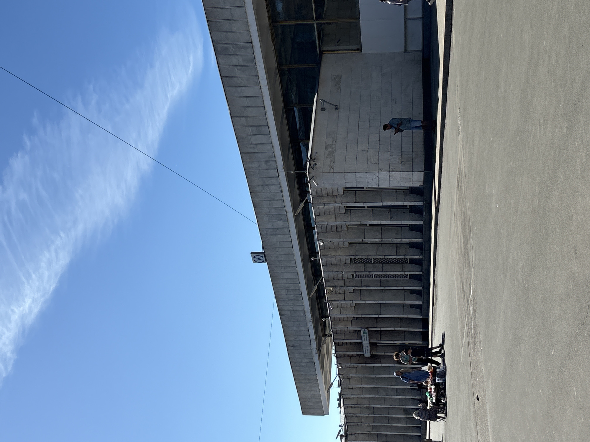 This image depicts a brutalist-style concrete building, an entrance pavilion to a Saint Petersburg Metro station in Russia, identified by the distinctive "M" logo on its roofline. The building features a large, flat, overhanging roof made of textured concrete panels, above a facade with dark, reflective windows. The lower section of the building is characterized by vertical concrete fins. An arrow sign near an entrance indicates "Метро / Metro".
The scene unfolds under a bright, clear blue sky with a few sparse, wispy clouds. A thin, dark cable or power line diagonally traverses the upper sky. In the foreground, a wide expanse of grey asphalt pavement is brightly illuminated by direct sunlight, casting sharp shadows.
On the left side of the frame, near the building's entrance, an elderly woman is seated behind a small, informal market stall displaying various goods. A woman in a long blue dress, another person with curly hair, and a child stand nearby, appearing to interact with or observe the stall. Further to the right, a man in a light blue shirt and shorts stands facing the building, absorbed in his mobile phone. The lower legs of another person are partially visible on the far right edge of the image.