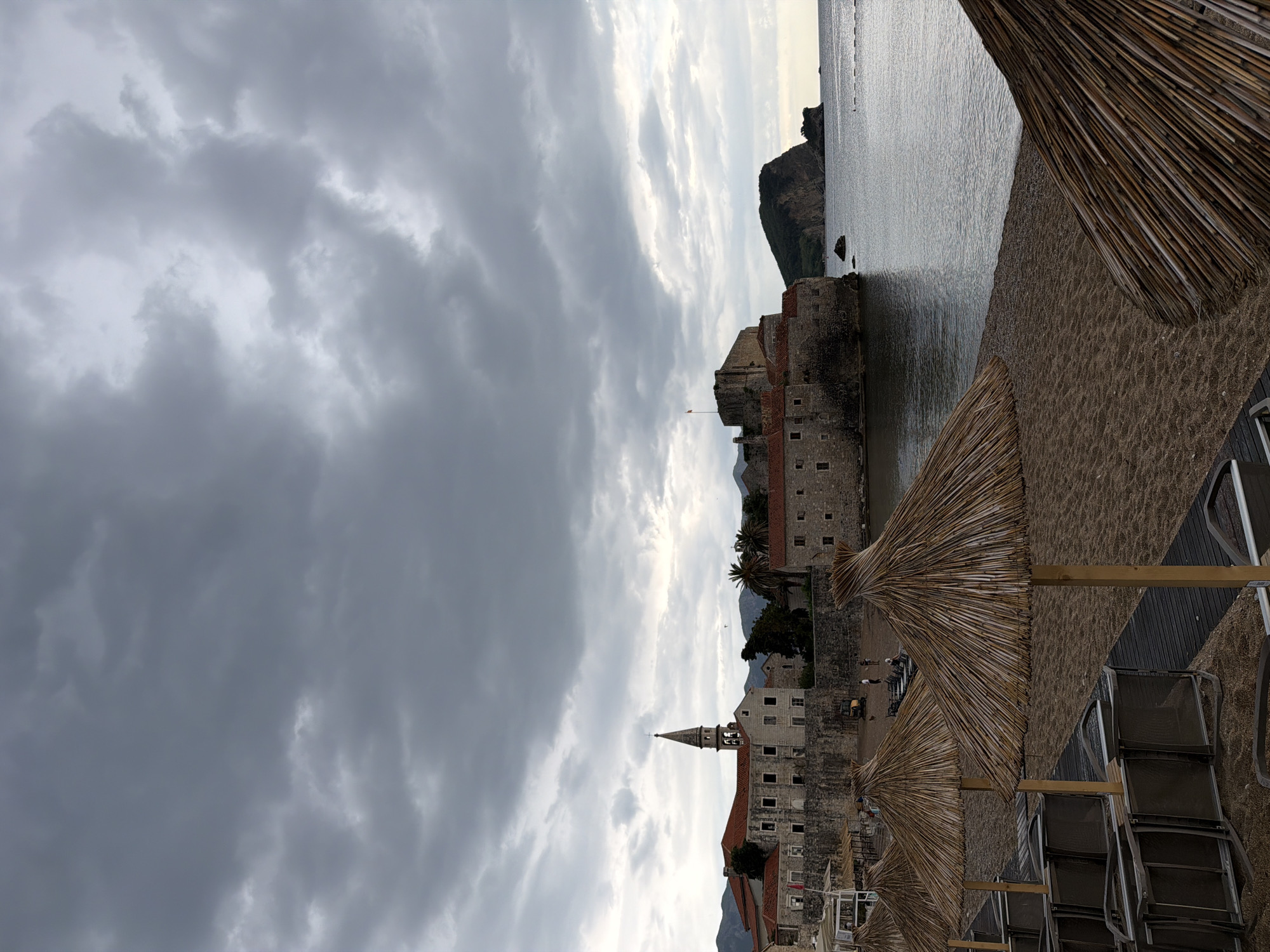The image depicts a cloudy day on the beach in Budva, Montenegro. The old town of Budva is visible with its historic stone buildings, including a church with a steeple. In the foreground are straw umbrellas and beach chairs. The sea is calm. A rocky islet is visible in the distance.