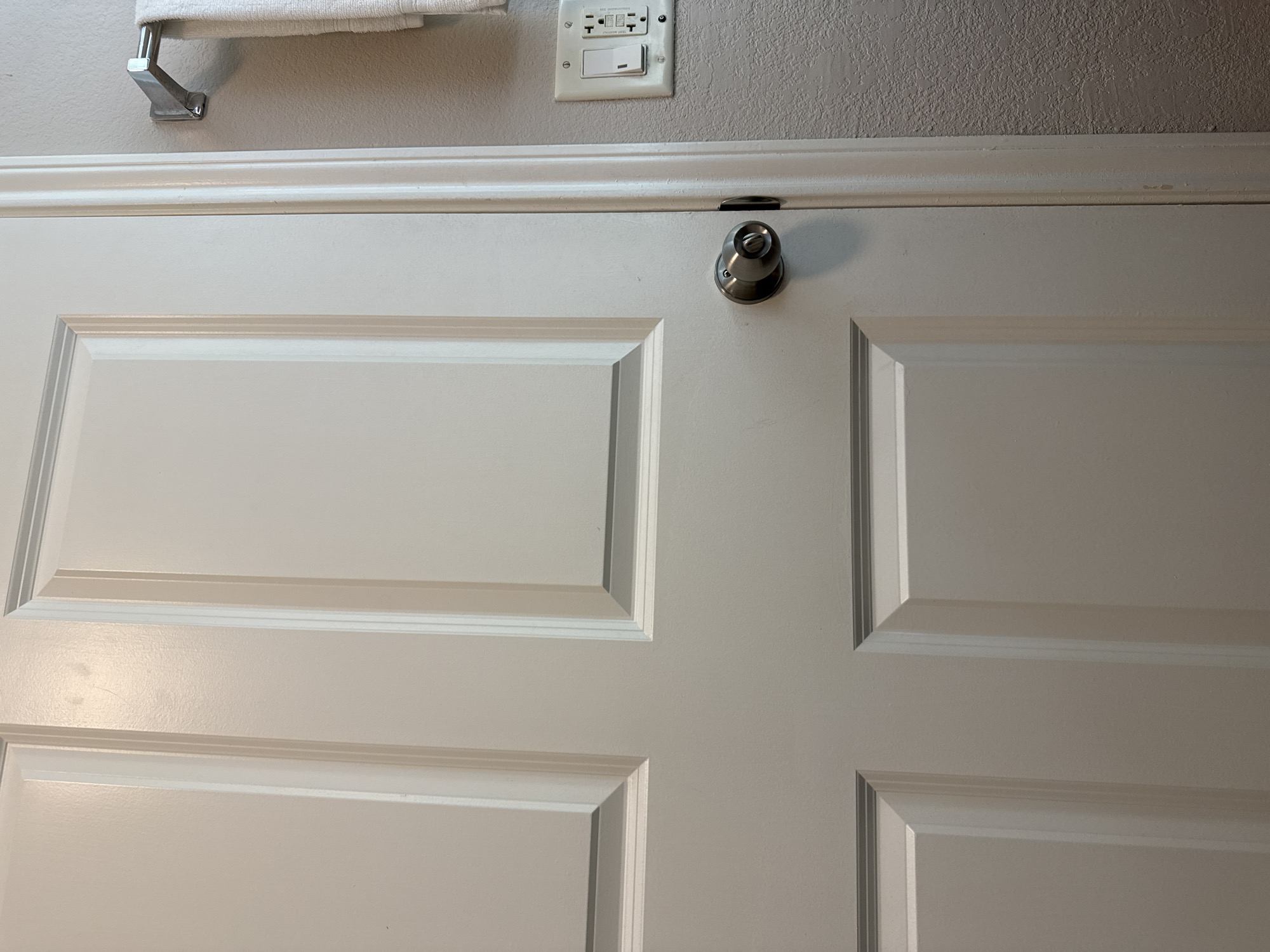 The image shows a close-up of a white, paneled door with a silver doorknob. Above the door is a section of wall with a towel rack and an electrical outlet. The wall is a light gray color. The scene appears to be indoors, likely a bathroom. The location is the city of San Antonio, United States of America. There are no people visible.