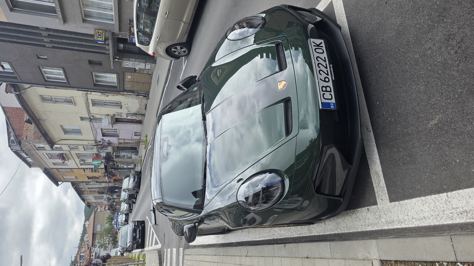 The image shows a green Porsche parked on a street in Blagoevgrad, Bulgaria. The car is parked in a designated parking space marked by white lines. The license plate number is CB 6222 OK. The street is lined with buildings of varying heights and colors. Other cars are parked along the street. The sky is overcast.