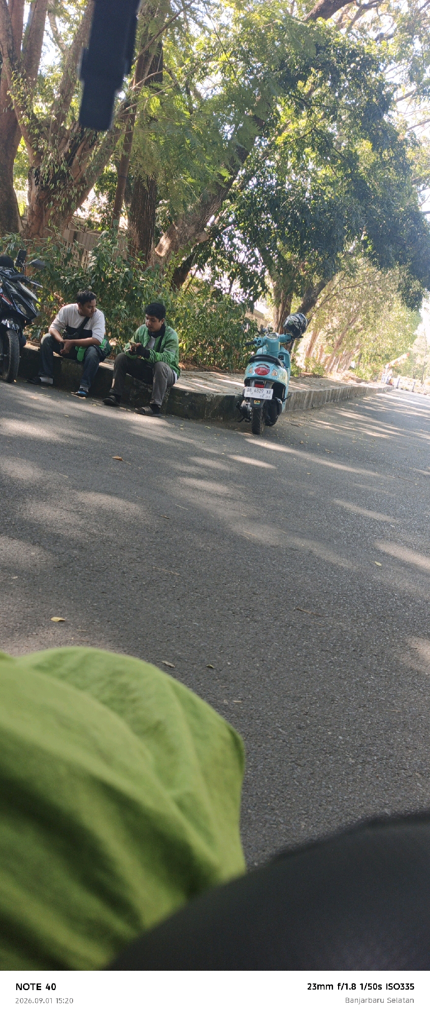 The image depicts an outdoor scene along a road in Banjarbaru, Indonesia. Two men are seated on a concrete curb next to a row of trees. The man on the left wears a white and grey long-sleeved shirt and dark pants, with a green bag slung over his shoulder. The man on the right is dressed in a green jacket and grey pants, and he is looking at his phone. To the right of the men, a light blue scooter is parked. Further down the road, other trees line the street, casting shadows on the asphalt. A motorcycle is partially visible on the far left of the image. The lower portion of the image shows a close-up of a crumpled green fabric, likely clothing, and the bottom edge of the frame. The timestamp and location information at the bottom of the image confirm it was taken on September 1, 2026, at 15:20 in Banjarbaru, South Kalimantan, Indonesia, with a 23mm lens at f/1.8 aperture and 1/50s shutter speed with an ISO of 335.