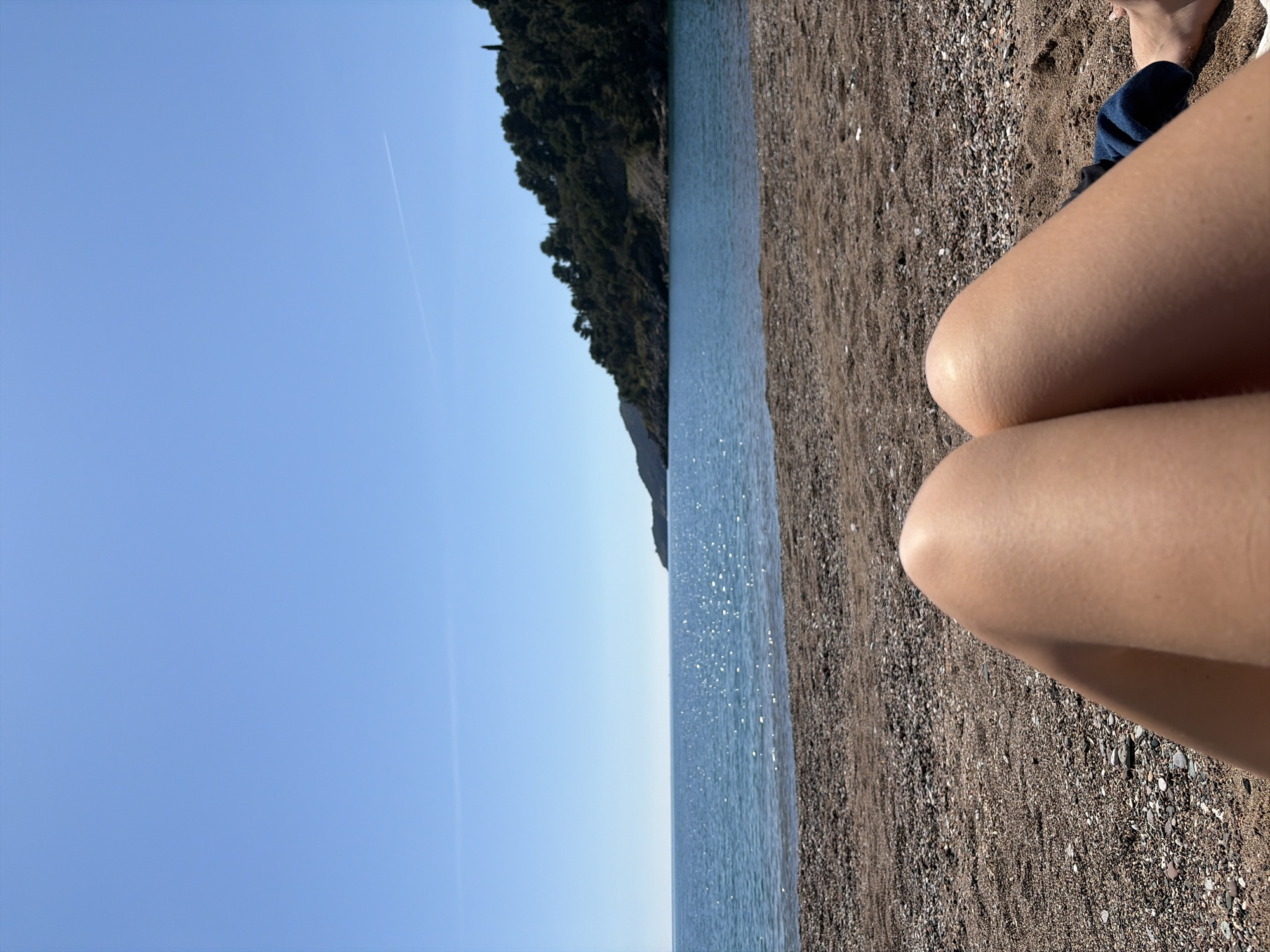 The image shows a beach scene. In the foreground are a pair of human legs on a pebble-strewn beach. A patch of blue fabric is visible near the feet. The middle ground consists of the sea and a tree-covered landmass in the distance. The sky is clear and blue with a faint contrail from an airplane. The location is in the city of Unknown, Unknown.