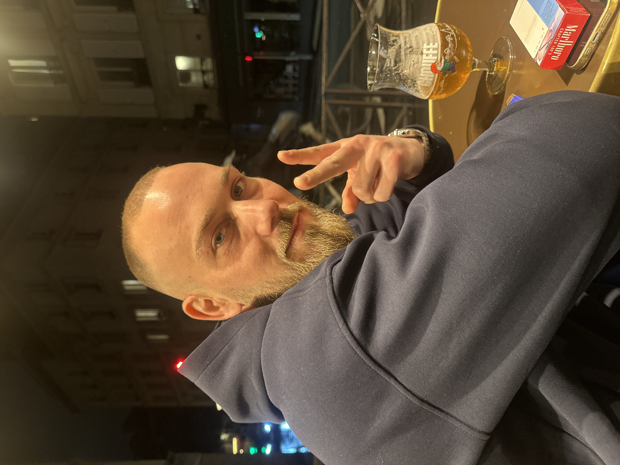 The image shows a Caucasian man with a shaved head, light blue eyes, and a blonde beard giving a peace sign. He is wearing a navy blue hooded sweatshirt. He appears to be sitting at an outdoor cafe table in Paris, France. A glass of beer, a pack of Marlboro cigarettes, and a cell phone are on the table. Streetlights and building facades are visible in the background.