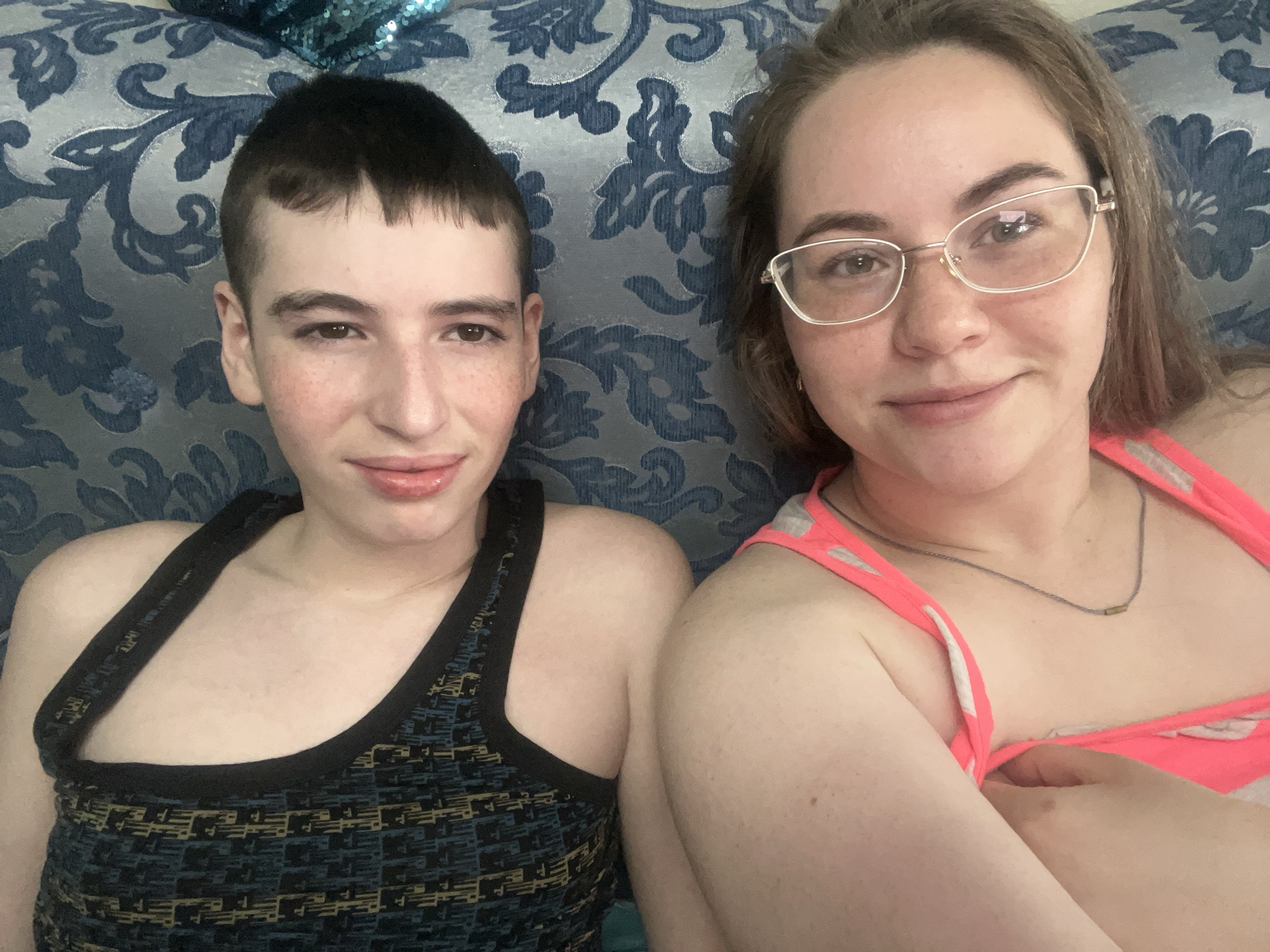 Two individuals are lying down in what appears to be an indoor environment. The person on the left has a very short, dark haircut and is wearing a patterned tank top. They have fair skin, with noticeable freckles around the cheeks and nose. The person on the right has light brown hair, and they wear glasses with a thin metal frame. They are wearing a light pink tank top with white trim and a thin necklace. The background is a blue and white patterned fabric, possibly a couch or bed covering. The image does not include any identifiable landmarks to determine the location in Chaikovsky, Russia. It primarily features a close-up of the individuals' faces and upper bodies.