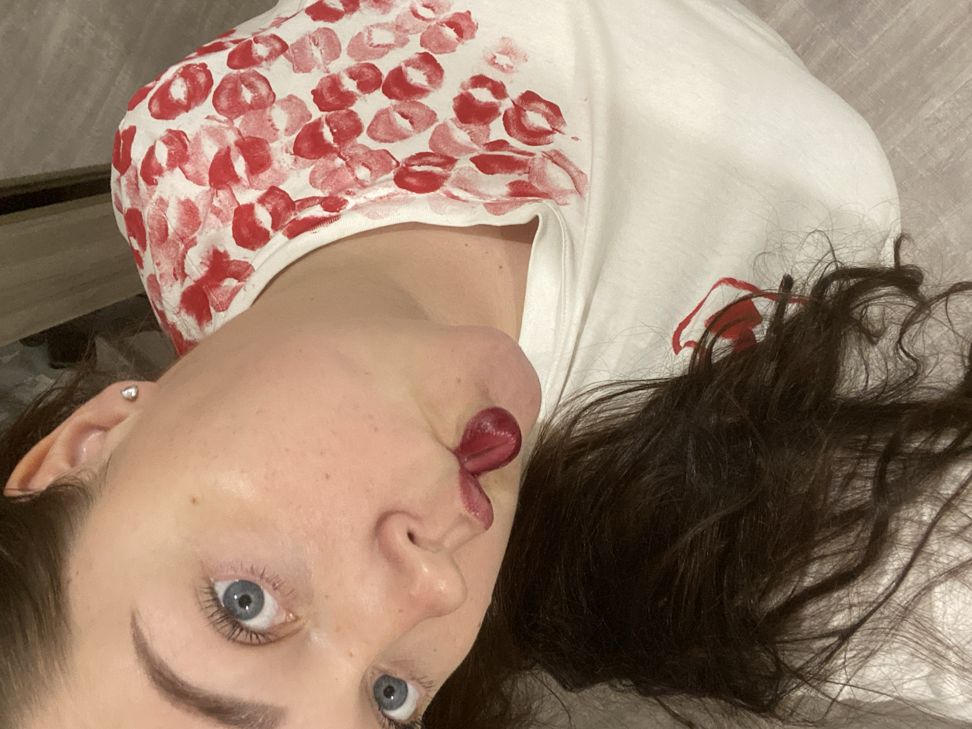 The image shows a woman lying down, possibly on a light-colored surface like a bed or floor. Her head is tilted to the side. She has fair skin, dark hair, and blue eyes. There is dark red lipstick smeared on her lips, with some smeared on her face as well. She is wearing a white t-shirt covered in red lipstick kiss marks on the shoulder and upper chest area. The exact location and association with Sochi, Russia, cannot be determined from the image alone.