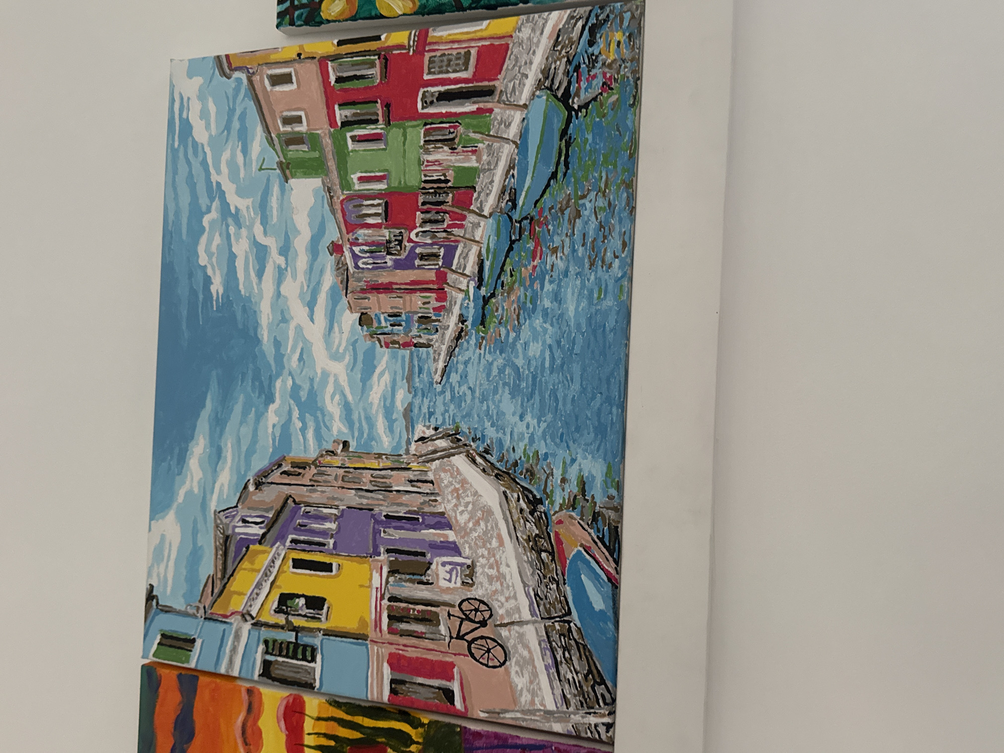 The image shows a painting depicting colorful buildings along a waterway under a blue, cloudy sky. The style is impressionistic with visible brushstrokes. The buildings feature various colors like red, green, purple, yellow, and blue, creating a vibrant scene. A bicycle is painted on the side of one of the buildings. The waterway reflects the colors of the buildings.
I am unable to identify the location in the city of Plovdiv, Bulgaria.