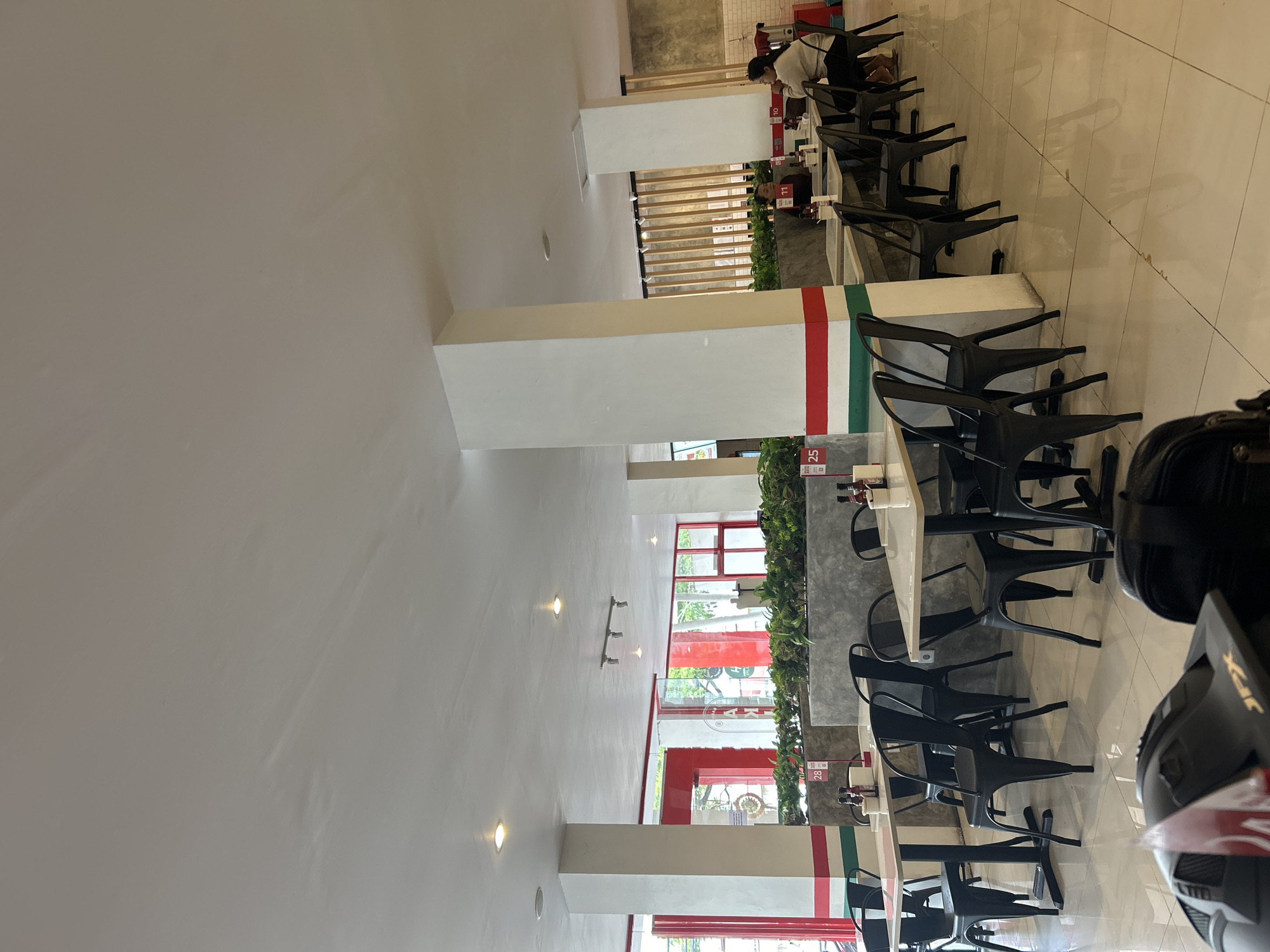 The image depicts the interior of a restaurant with multiple tables and chairs. The tables are small and square, with white tops and black legs. They are set with black chairs. The restaurant has a modern and clean aesthetic, with white walls and a tiled floor. There are several potted plants placed around the restaurant, adding a touch of greenery. In the background, there are large windows that allow natural light to enter the space. Some of the tables have bottles and containers on them, suggesting that food and drinks are served. There are a few people visible in the image, one person is sitting at a table in the background and another person is standing near a counter. The overall impression is that of a casual dining establishment.