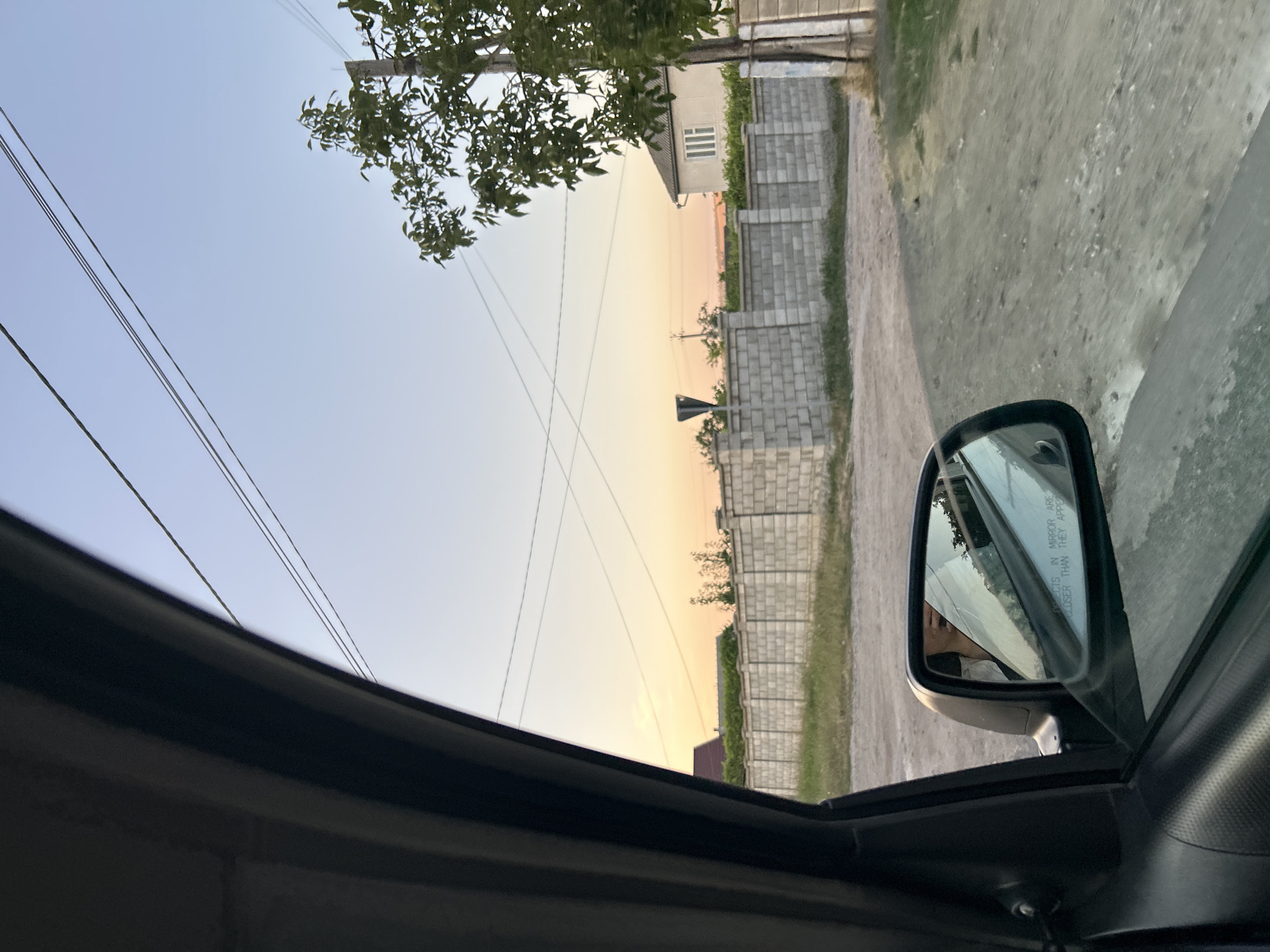 The image shows a view from inside a car, looking out through the side window and the side-view mirror. The scene appears to be a rural or semi-rural area, possibly in the city of Abaclia, Moldova. A dirt road is visible in the foreground, with grass and some loose gravel. Beyond the road, there is a low, grey brick wall. Behind the wall, a building with a light-colored facade and a dark roof is partially visible. A tree with green leaves stands to the right of the building. Several power lines are strung across the pale blue sky, which is transitioning to a warm orange and yellow hue near the horizon, suggesting either sunrise or sunset. The side-view mirror reflects the interior of the car, including a person's face and upper body, holding a phone. Text on the mirror reads "OBJECTS IN MIRROR ARE CLOSER THAN THEY APPEAR."