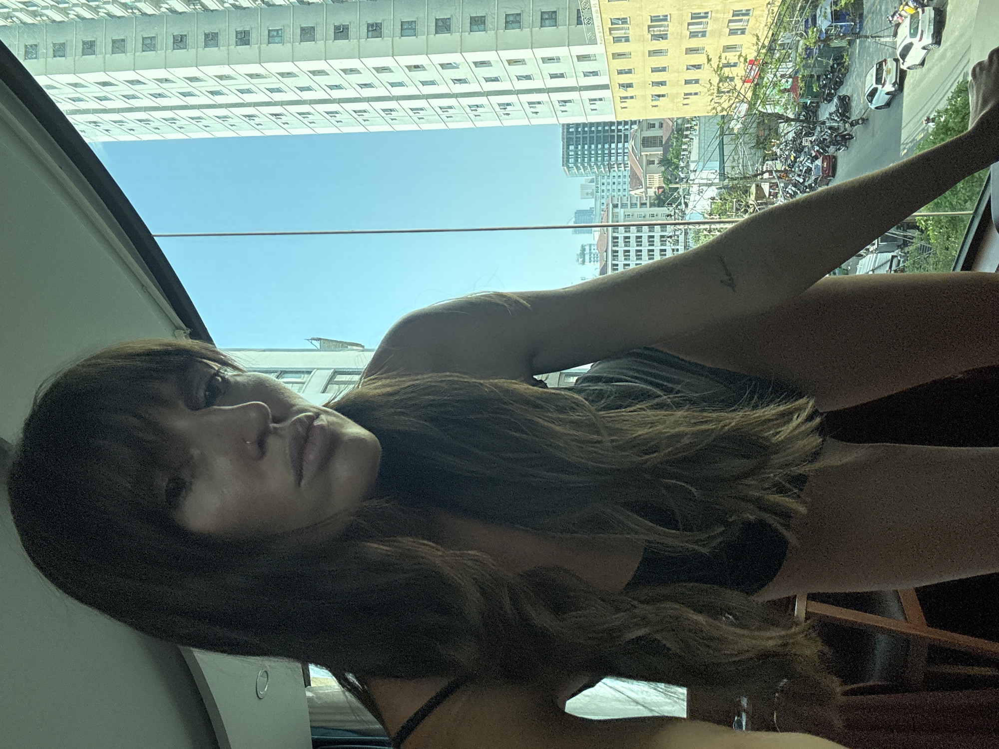 A woman with long dark hair and bangs lies facing the camera, with a view through a window of a densely populated urban environment. The location appears to be Đà Nẵng, Vietnam, based on architectural styles and the prevalence of motorbikes on the street below. The buildings visible through the window range in height from a few stories to high-rise structures. There are parked motorbikes and cars on the street. The sky is clear and blue. The woman is wearing what appears to be a black top. A tattoo is visible on her arm.