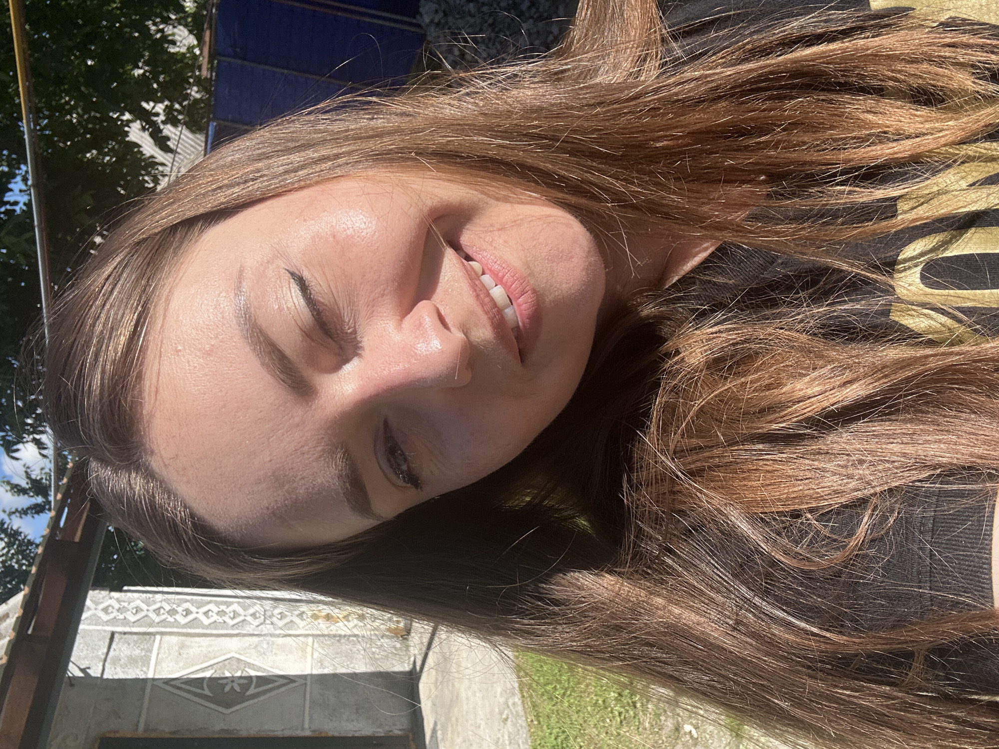 In Bila Tserkva, Ukraine, a young woman with long, brown hair smiles at the camera in a close-up selfie. Her left eye is partially closed, and she is looking slightly to the right of the lens. Her skin appears smooth and sun-kissed, with a hint of natural blush on her cheeks. The sunlight casts a warm glow on her face, highlighting subtle contours and a few scattered freckles. Her lips are parted in a gentle smile, revealing pearly white teeth. She is wearing a dark t-shirt with a faint, golden logo visible on the front.
The background is softly blurred, but suggests an outdoor setting. To the left, a light-colored, textured wall with a decorative pattern is visible, possibly part of a building. Behind her, trees with lush green foliage are discernible, along with a hint of a blue structure. The overall atmosphere is bright and casual, implying a pleasant day outdoors.