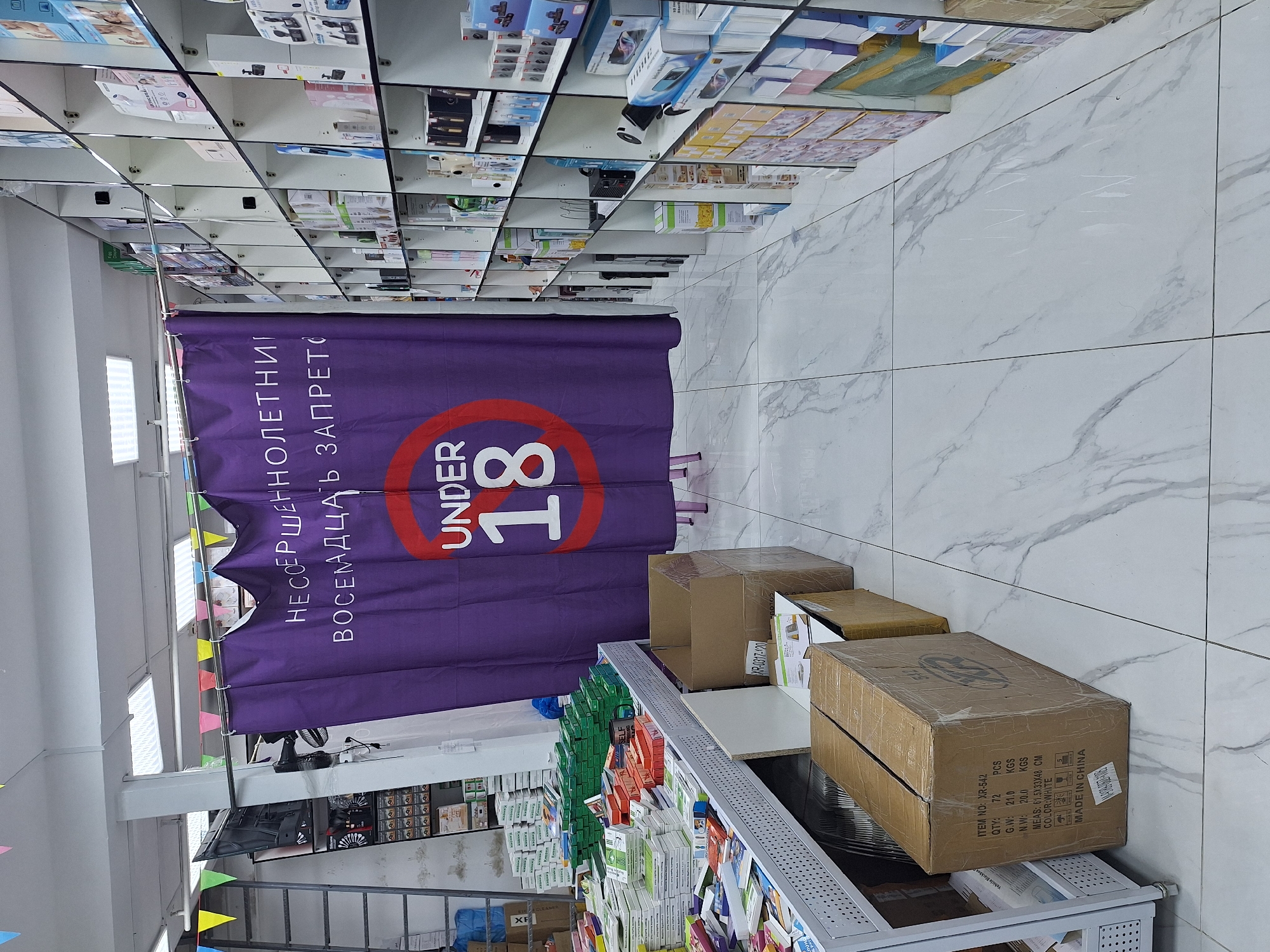 Here is a detailed description of the image:
The image shows the interior of a shop, possibly in Moscow, Russia, judging by the Russian text. A purple curtain hangs, displaying a red circle with "UNDER 18" written inside, and above it, the Russian text "НЕСОЕРШЕННОЛЕТНИМ ВОСЕМНАДЦАТЬ ЗАПРЕТО," which translates to "Underage eighteen prohibited." The curtain is decorated with colorful pennant flags.
In the foreground, a metal shelf unit is stocked with various items, including boxes of medicines. Brown cardboard boxes are placed on the shelves and the floor near the curtain. The floor is covered with light-colored, marble-like tiles.
Above the scene, the shop's ceiling has a grid-like structure, with shelves full of merchandise and boxes. The lighting is bright, illuminating the shop interior. The scene appears to be taken inside a retail establishment.