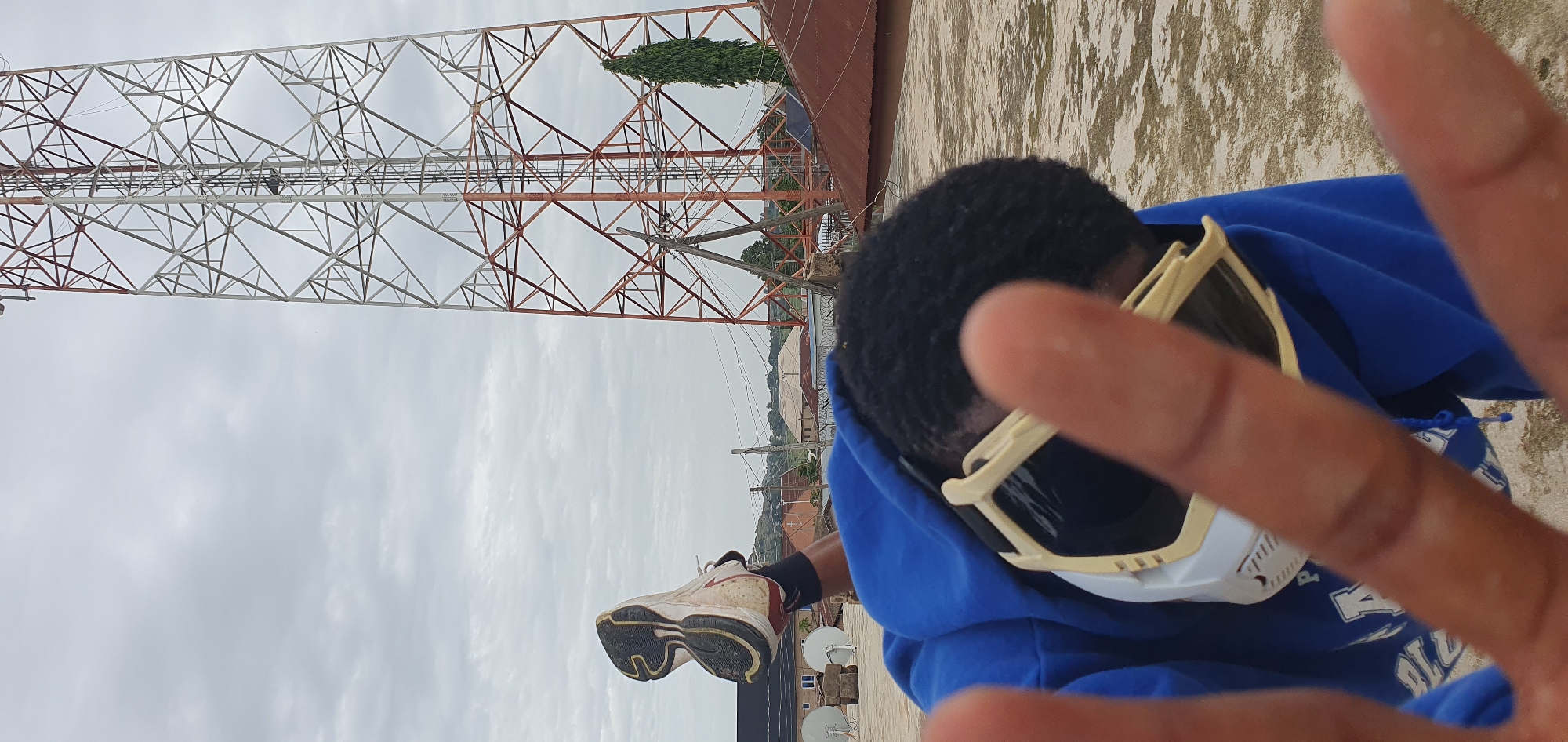 A person wearing a blue hoodie and protective goggles is shown from the chest up, with their hands in the foreground, obscuring their face. Their right leg is extended upwards, with a white and black sneaker visible. In the background, a large, red and white lattice tower stands tall against an overcast sky. The scene appears to be on a rooftop in Anguldi, Nigeria, with various buildings and a tall green tree visible in the distance. The ground is a flat, textured surface, possibly concrete or asphalt.