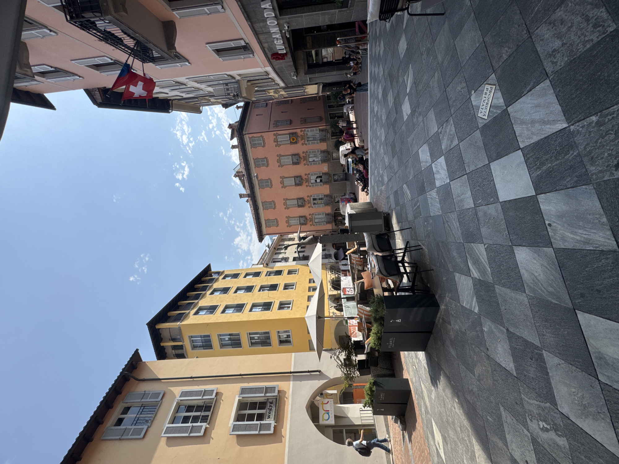The image captures a sunlit outdoor plaza in Lugano, Switzerland, featuring a distinctive grey, diamond-patterned stone pavement. On the left, an orange building displays white-shuttered windows and an arched entrance labeled "art COMPUTER." Next to it, a tall yellow building showcases multiple floors with dark-framed windows. Across the plaza, pink and coral buildings line the street, adorned with more white-shuttered windows and balconies. Swiss and red flags are visible on a balcony of one of the pink buildings. Outdoor seating areas with tables, chairs, and umbrellas are set up in front of cafes and restaurants, with several patrons seated or walking. People are seen walking along the sidewalk and in the plaza, some with backpacks. Planters containing greenery are placed strategically. The sky is a clear blue with a few wispy clouds. In the foreground, a metallic plaque embedded in the pavement reads "PROPRIETA PRIVATA."