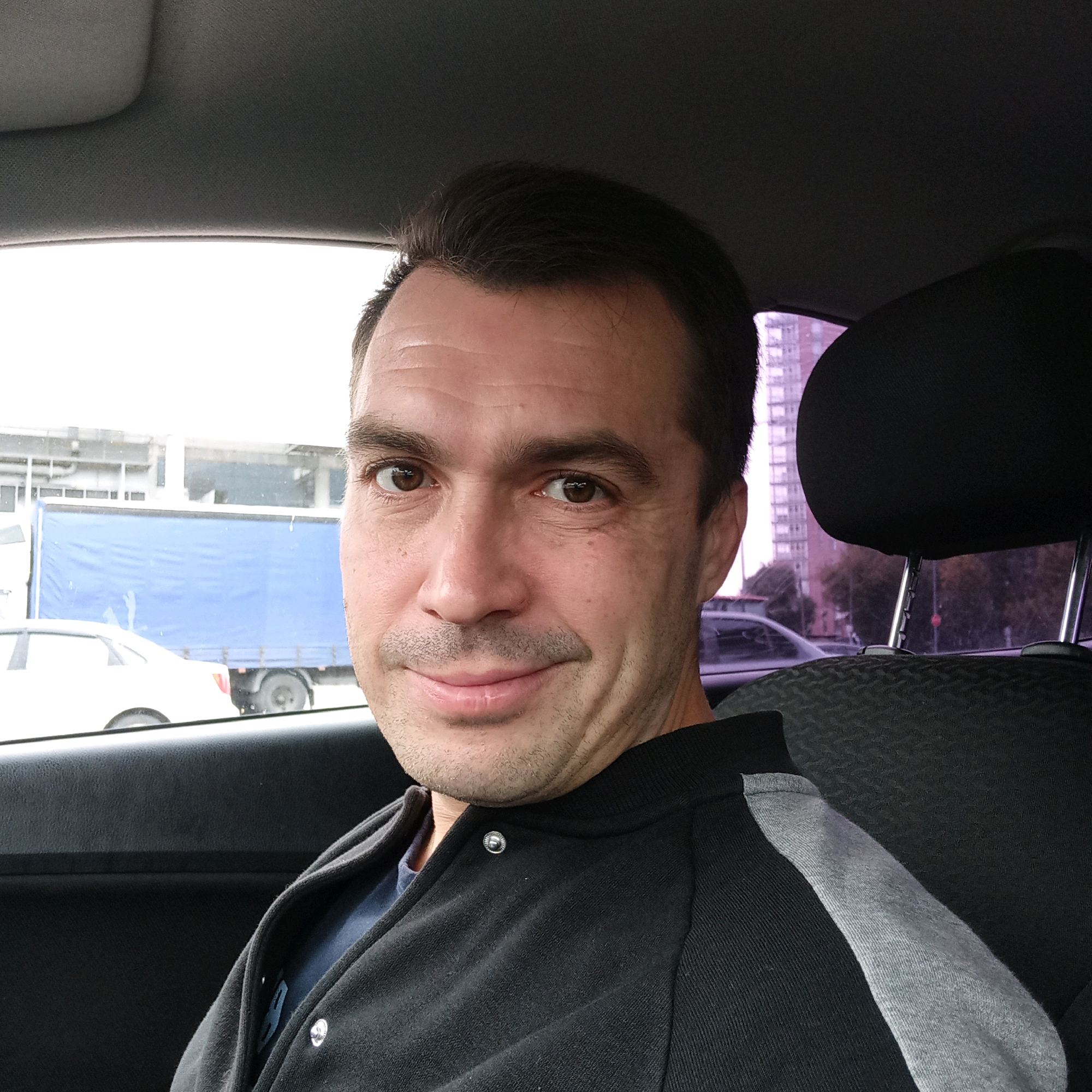 A man is taking a selfie in the passenger seat of a car. He is smiling and looking at the camera. The car is parked on a street with a large blue truck and other vehicles in the background. The man is wearing a black and gray jacket. The scene appears to be a street in Nizhny Novgorod, Russia.