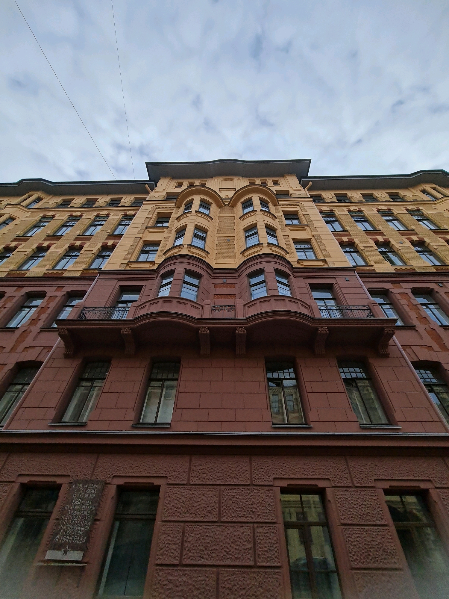 This is a low-angle, upward shot of the exterior of a multi-story building in Saint Petersburg, Russia. The building has a distinct architectural style with a tan-colored upper section and a reddish-brown lower section. The tan section features numerous evenly spaced windows and decorative cornices. The reddish-brown section is textured and has fewer, larger windows, some with ornate balconies. A plaque with Cyrillic text and a red star is visible on the lower left portion of the building. The sky above is overcast with scattered clouds, and two thin wires are stretched across the upper part of the frame. The perspective creates a dramatic effect, emphasizing the height and grandeur of the building.