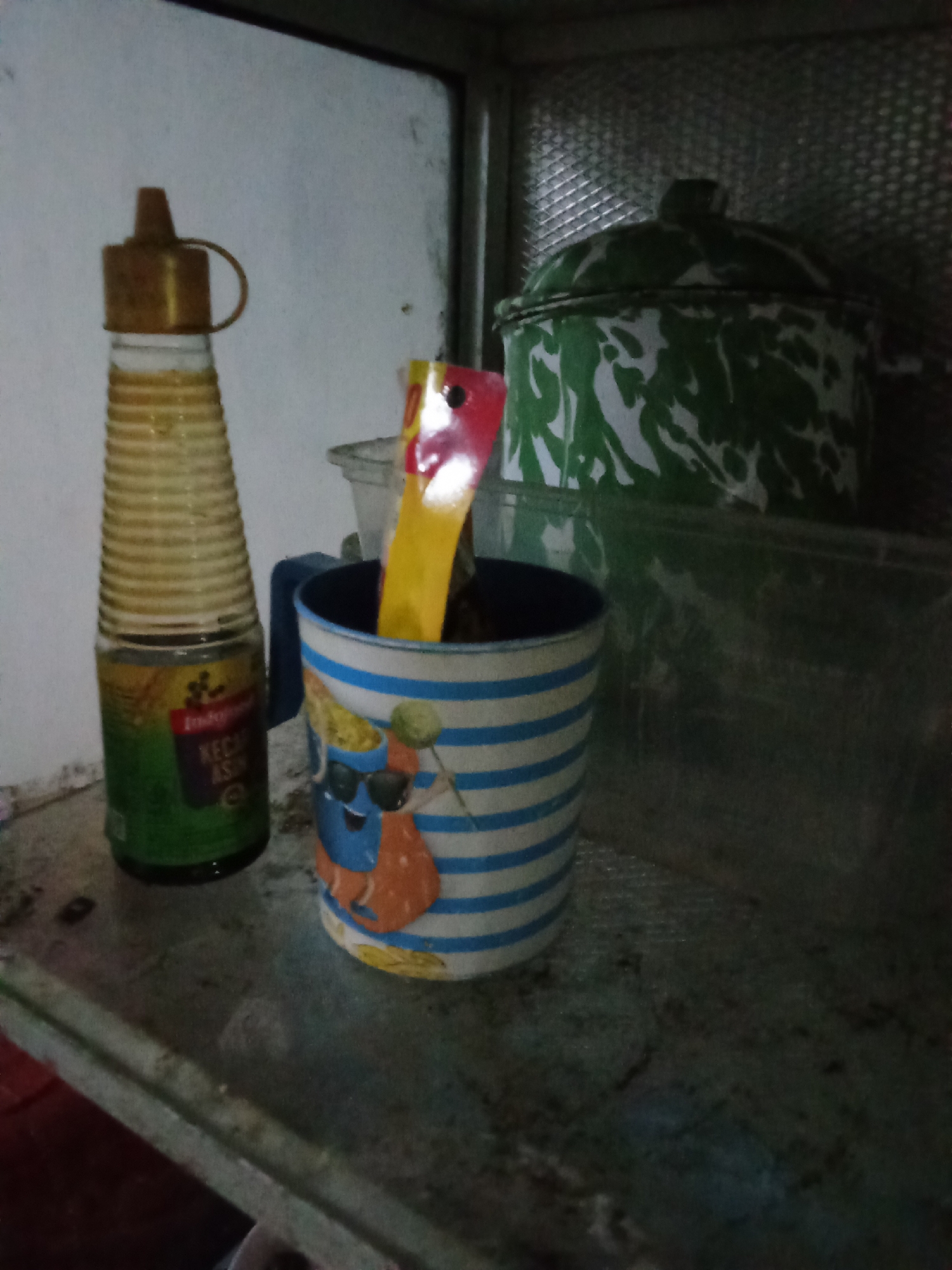 The image depicts a shelf in what appears to be a kitchen or pantry, possibly in the city of Pemalang, Indonesia. On the shelf, a tall, ribbed glass bottle with a gold cap is visible on the left, containing a light-colored liquid and labeled with text including "Indofood" and "Kecap Asin". To the right of the bottle, a blue and white striped mug holds a bright yellow and red object, possibly a spoon or a utensil handle. Behind the mug and bottle, a large, green and white patterned pot with a lid is situated, and further back, a transparent plastic container can be seen. The shelf itself is dusty and shows signs of wear. The lighting is dim, casting shadows and creating a slightly muted atmosphere.