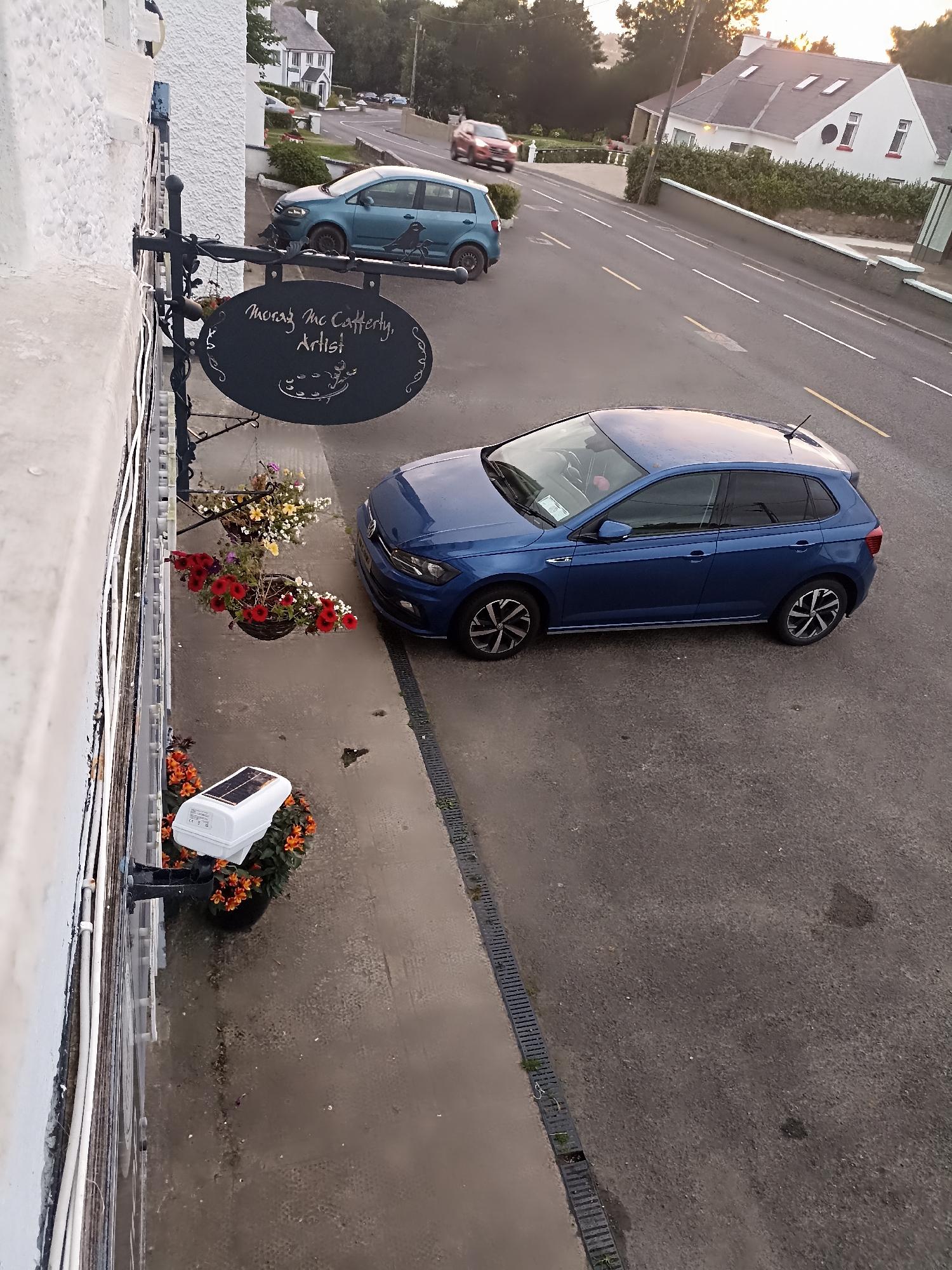 The image depicts a street scene in Annagry, Ireland, featuring a blue Volkswagen Polo parked on the right side of the road. A light-colored building with white textured walls and some pipes is visible on the left. Hanging from the building is a dark oval sign with white lettering that reads "Morag McCafferty, Artist". Below the sign, there are two hanging baskets filled with red and white flowers. Further down, a white solar-powered security light is mounted on the wall, with orange flowers planted around its base. In the background, other buildings and a road with cars can be seen, bathed in the soft light of what appears to be either sunrise or sunset.
