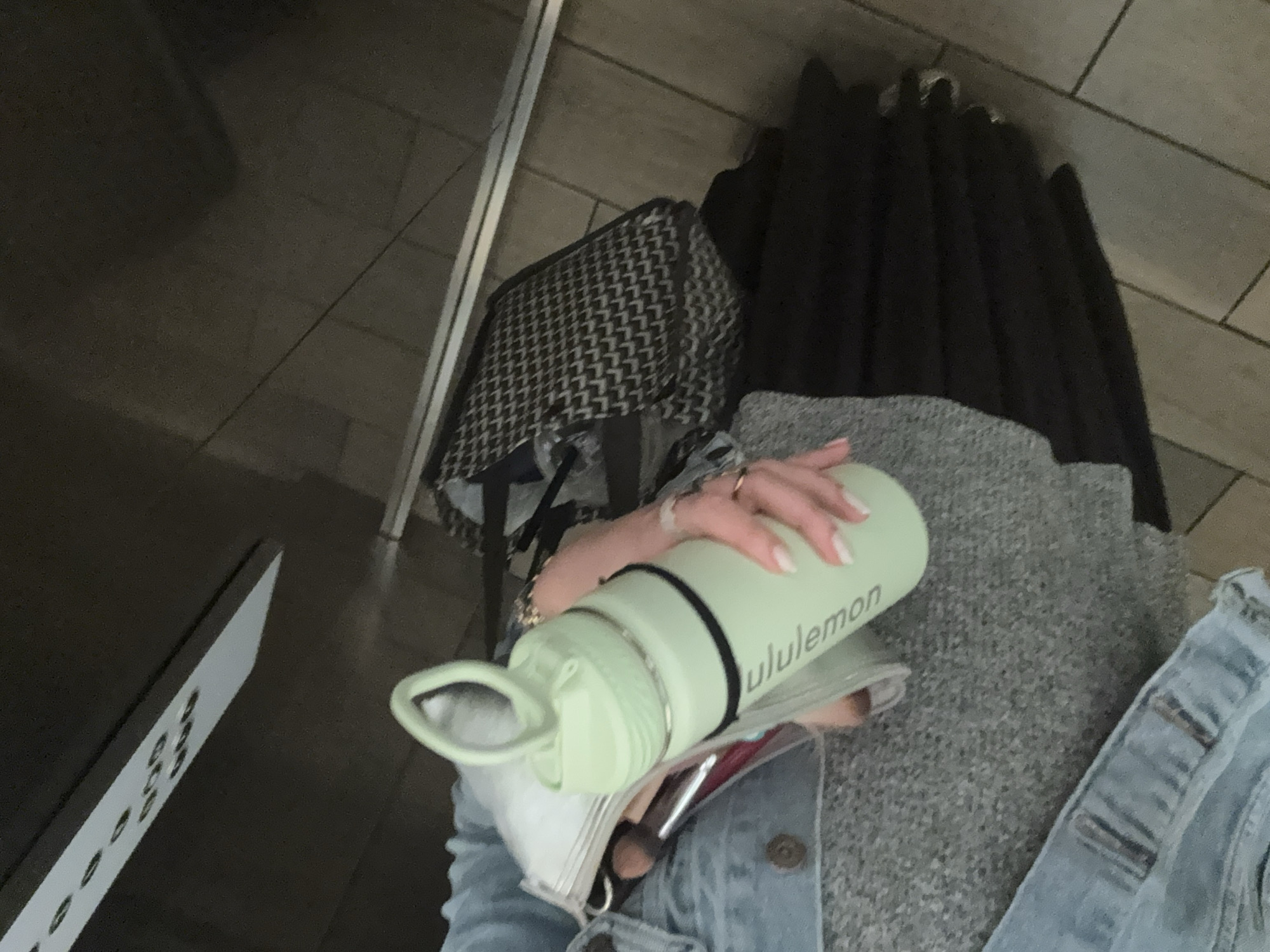 The image shows a person's lap. The person is wearing a light denim jacket and gray pants. They are holding a light green water bottle with the Lululemon logo. The water bottle is on top of a white bag with makeup brushes inside. Behind the person is a black bag and a row of black coats. The location is Mexico City, Mexico. The floor is tiled. There is a white object on the left side of the image that is likely part of a table or cabinet.