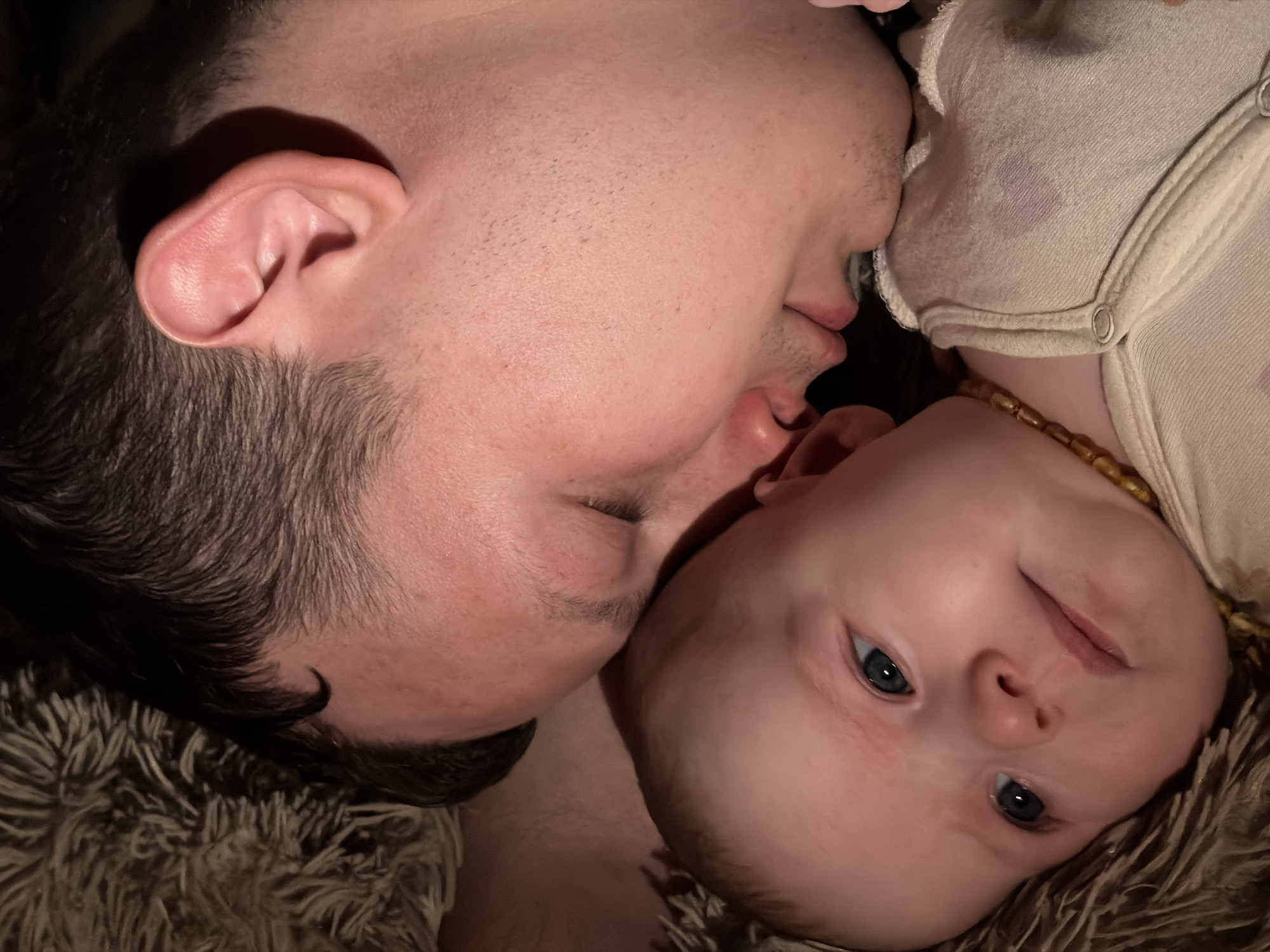 An intimate portrait, captured in Chaikovsky, Russia, features an adult and a baby lying close together on a soft, textured, brownish blanket. The adult, positioned on the left, has short, dark hair and noticeable stubble on their cheek and jawline. Their eyes are closed or cast downwards, and their mouth is slightly open, conveying a sense of peace or tender interaction.
The baby, on the right, has fair skin, bright blue eyes, and is looking directly at the camera with an alert, curious, and subtly smiling expression. A beaded necklace, likely amber, is visible around the baby's neck, and they are wearing a light-colored, possibly cream or white, onesie with visible snaps. A faint purple stain is present on the baby's clothing near the neckline.
The lighting is soft and warm, suggesting an indoor, cozy setting, possibly in the evening. The overall mood of the image is one of profound affection, serenity, and connection between the adult and the infant. There is no visible text.