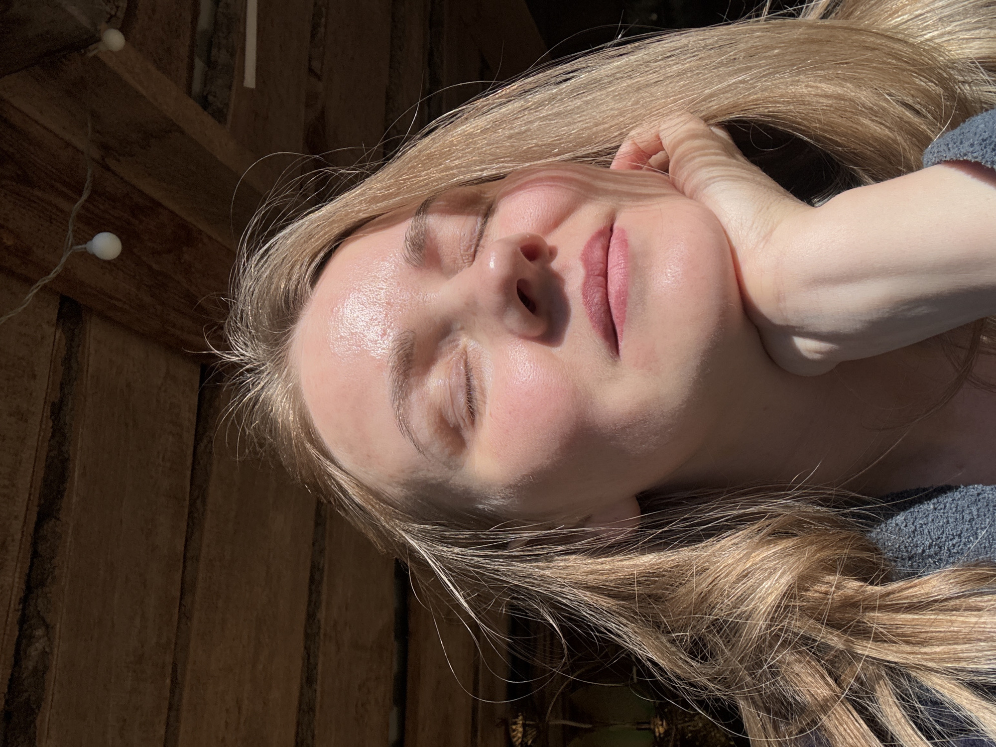 A fair-skinned woman with closed eyes is depicted in an indoor setting, likely in Chaikovsky, Russia, based on the query context. She is resting her face on her hand, and the sunlight casts shadows on her face and blonde hair. The background features a wooden wall and a string of lights with spherical bulbs. The woman is wearing a gray garment with a knitted texture.