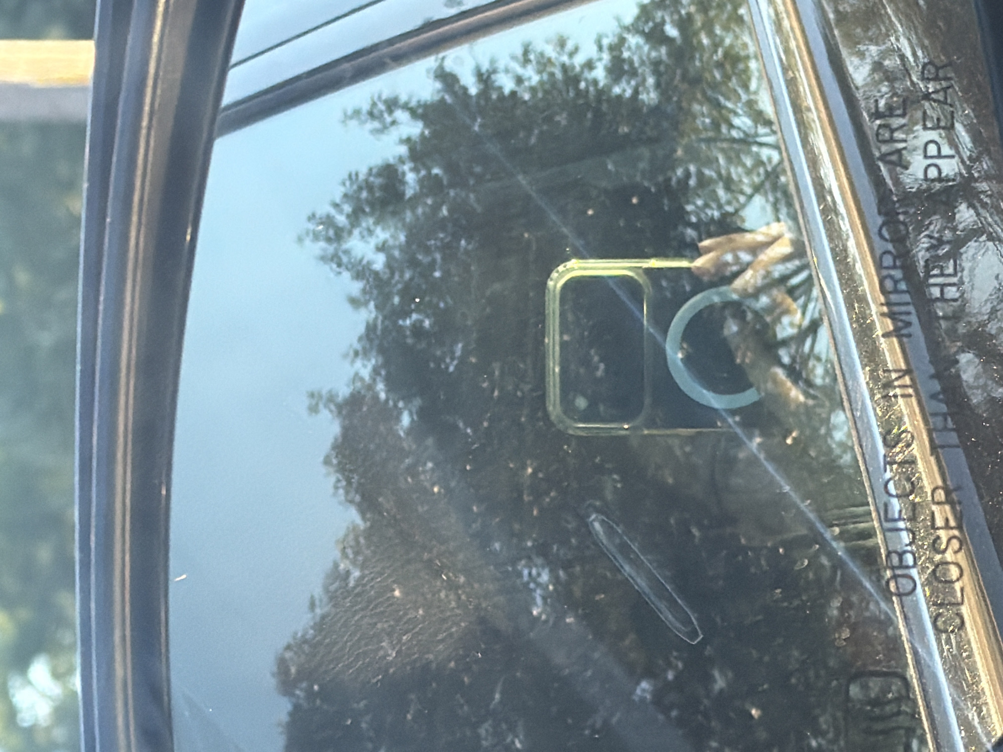 The image shows a close-up of a car's side mirror, reflecting trees and sky. Text is visible on the mirror: "OBJECTS IN MIRROR ARE CLOSER THAN THEY APPEAR". A smartphone with a green case and a ring holder is also reflected in the mirror. The overall scene suggests a car parked outdoors, possibly in a suburban or rural setting, given the reflection of trees. Without further context, it is not possible to pinpoint the exact location in Riverhead, United States of America.