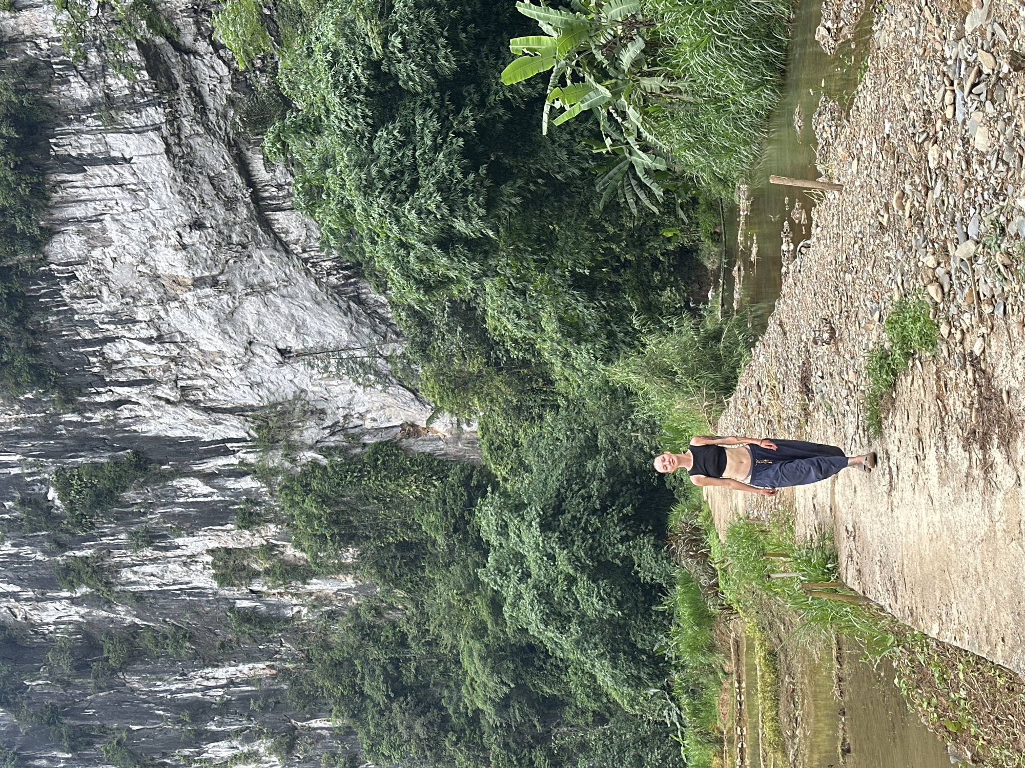 A woman walks down a dirt path next to rice paddies and a stream in Pù Luông Commune, Vietnam. The woman is wearing a black tank top and navy blue pants. She has short, blonde hair and is bald on the sides of her head. She is wearing sandals and has tattoos on her arms. The path is made of dirt and gravel, and there are green trees and a large cliff face in the background. The stream is shallow and clear, with rocks and pebbles on the bottom. The rice paddies are filled with water and are bordered by wooden stakes and green grass. The sky is overcast and the light is diffused.