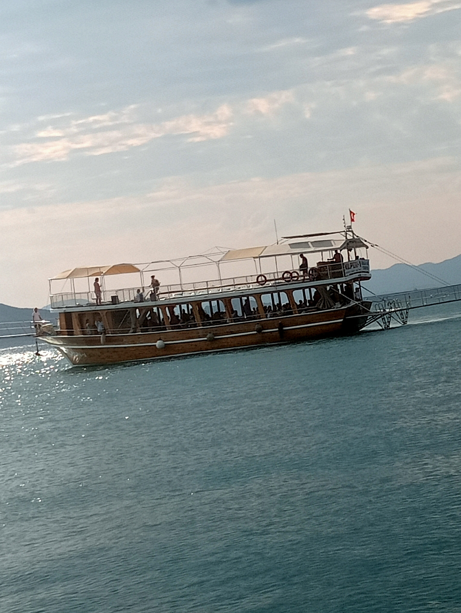 A wooden boat with multiple decks is sailing on the water. The boat is filled with passengers and has awnings for shade. The water is a calm, turquoise color. In the background, there are hills or mountains. The sky is partly cloudy with a hint of sunlight. A Turkish flag is visible on the boat. The boat appears to be on the Aegean Sea or Mediterranean Sea, common in Fethiye, Turkey, a popular tourist destination known for its boat tours and beautiful coastline.