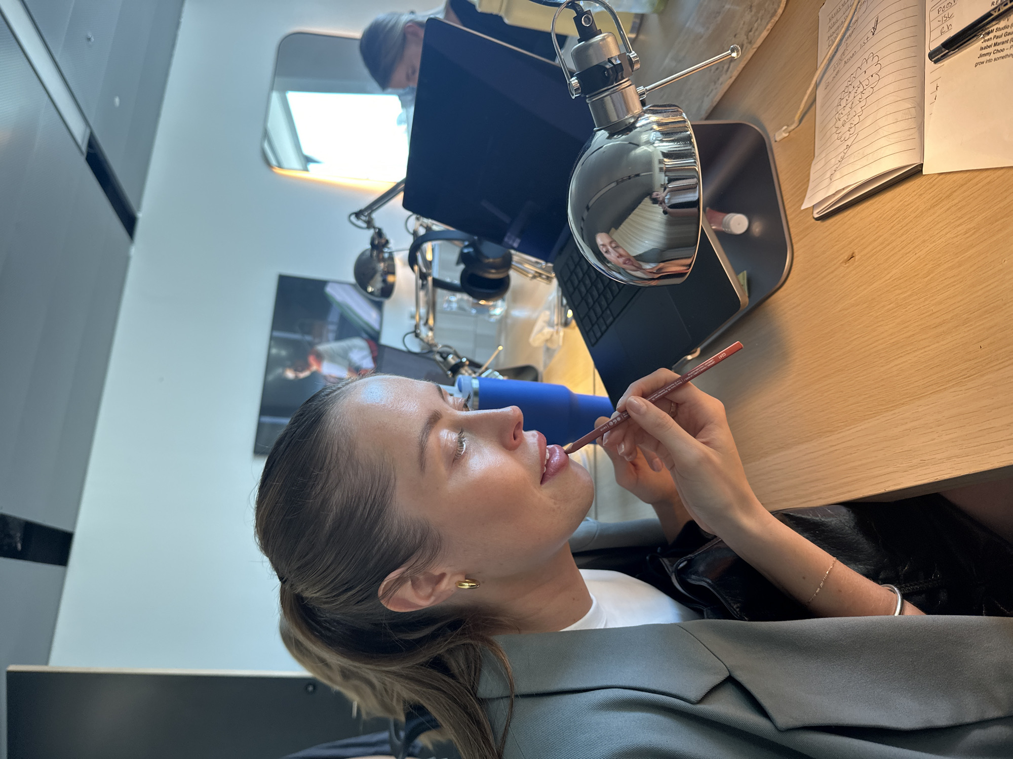 A woman is applying lip liner in front of a desk with a laptop, microphone, notebook, and blue water bottle. She is wearing a gray blazer and a white shirt, with gold earrings and a bracelet visible. The scene appears to be indoors, possibly a studio or office. The background includes a mirror, a framed picture on the wall, and office equipment. The woman is looking at her reflection in the microphone's surface as she applies the lip liner. There is no information in the image to identify the location as London, United Kingdom.