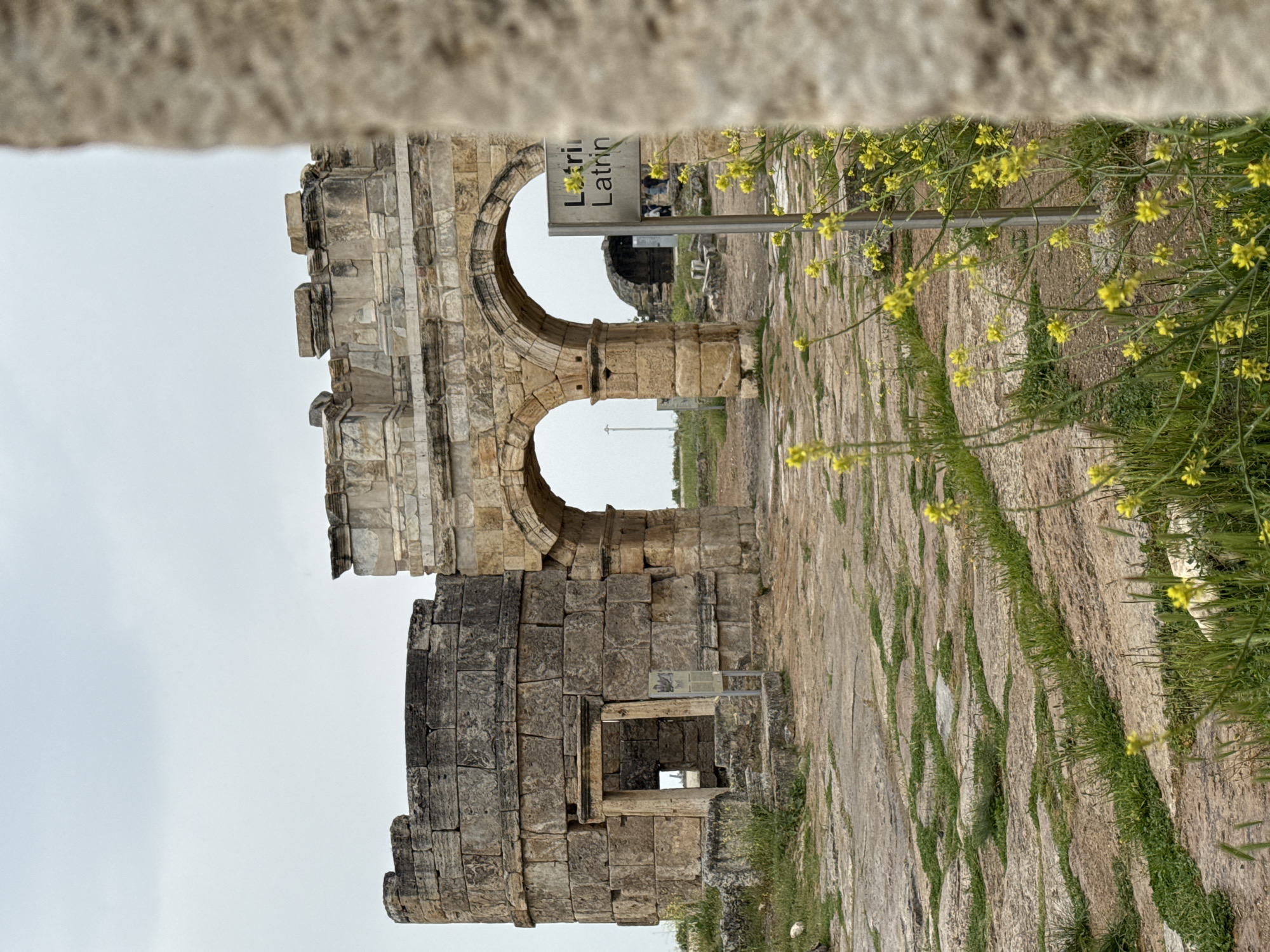 The image shows the ruins of Latrina, a public toilet in the ancient city of Hierapolis, now Pamukkale, Turkey. The structure is made of stone and features two arches. A sign with the word "Latrina" in both English and Turkish is visible near the arch. The ground is paved with stone, with grass and yellow wildflowers growing in the cracks. A section of the building's lower level features a window or opening. The sky is overcast and the weather is cloudy.