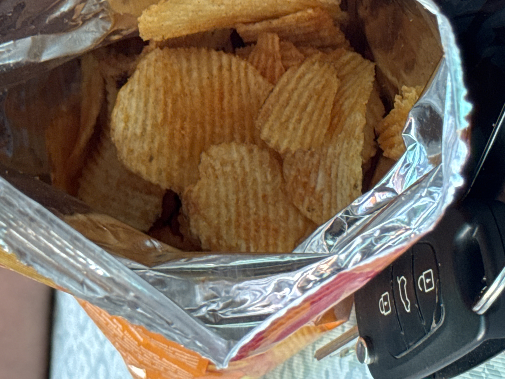 This is a close-up, indoor, eye-level shot of a partially opened bag of ridged potato chips. The bag is metallic and orange, with the chips spilling out. A black car key fob with three buttons is visible in the lower-left corner of the frame. The background is out of focus and appears to be a textured surface, possibly a tablecloth. There are no people or animals visible in the image. The scene is consistent with an outdoor setting on a warm day, given the apparent sunlight and casual placement of the items. The car key fob suggests the owner may have been driving or is about to drive. The context of Pribrezhnyi, Russia, indicates a potentially coastal environment, though the image itself does not provide any direct visual cues to confirm this. The overall impression is one of a brief, spontaneous pause, possibly a snack break during a journey.