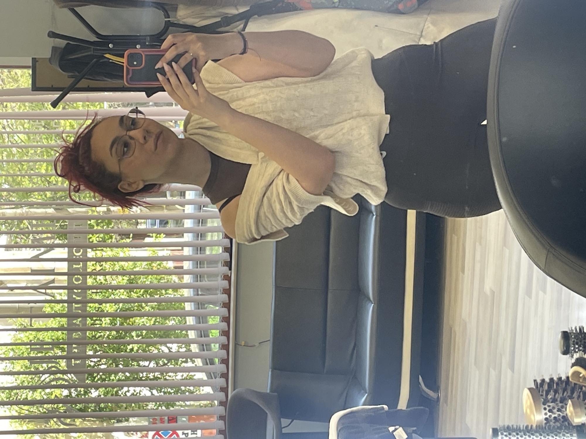 The image is a mirror selfie, originally captured sideways, featuring a young woman in an indoor setting. The woman, positioned on the left, has reddish hair tied up in a messy style and wears round eyeglasses. She is dressed in a dark brown top, black leggings, and a light-colored, possibly off-white or beige, knitted cardigan draped over her shoulders. She holds a smartphone with an orange-red protective case in her hands, capturing her reflection. Her expression appears neutral and composed.
The background, reflected in the mirror, reveals an interior space with a large window fitted with horizontal white blinds on the left. Through the blinds, bright natural light illuminates lush green trees and a glimpse of what appears to be a building or street scene outside, indicating a daytime setting. Visible outside the window, through the blinds, is inverted vertical text that partially reads "PRIYOLOL" or "PRIOL", and a red and white triangular sign with some faint letters like "H".
Within the room, to the right of the window, a black metal stand is visible. Further into the space, a large black leather-like sofa or bench is situated against a light-colored wall. A dark grey or black chair is in the mid-ground. On the far right, a large, dark, round object, possibly an exercise ball, is partially obscured. In the right foreground, several hair styling brushes or rollers with metallic and black components are arranged. The flooring is a light-colored wood. The environment suggests a commercial space, possibly a salon or studio, in Pleven, Bulgaria, based on the external text.
