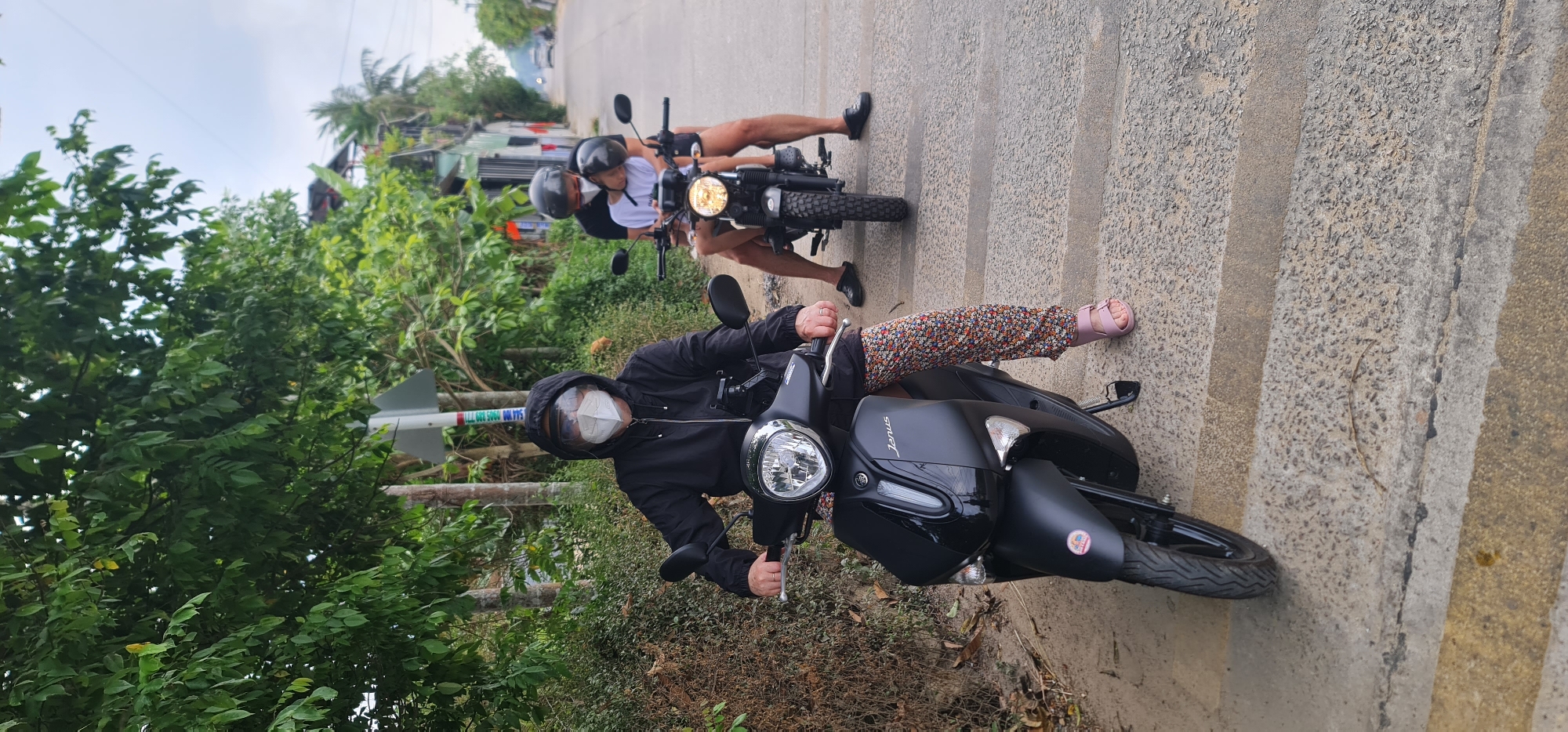 The image depicts a paved road scene in Hội An Ward, Vietnam. Two motorcycles are present. The motorcycle in the foreground is a black Janus model, carrying a person wearing a black hooded jacket, patterned pants, and pink sandals. The person is wearing a face mask. Behind the Janus motorcycle is a smaller motorcycle with two people wearing helmets. The background includes trees, buildings, and a utility pole with directional signs.