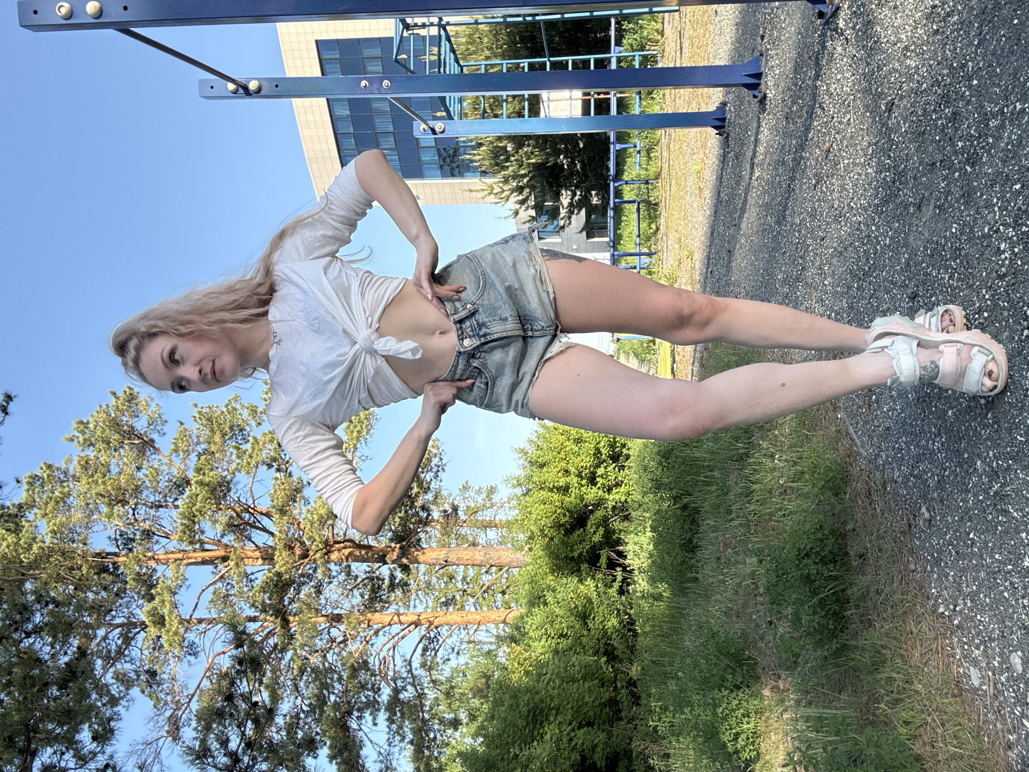 A blonde woman poses in an outdoor setting. She wears a tied white top and denim shorts, with bulky sandals on her feet. She stands on a gravel path near green grass and trees. In the background, there's a blue playground structure and a building. The scene appears to be in Прикамский, Russia.